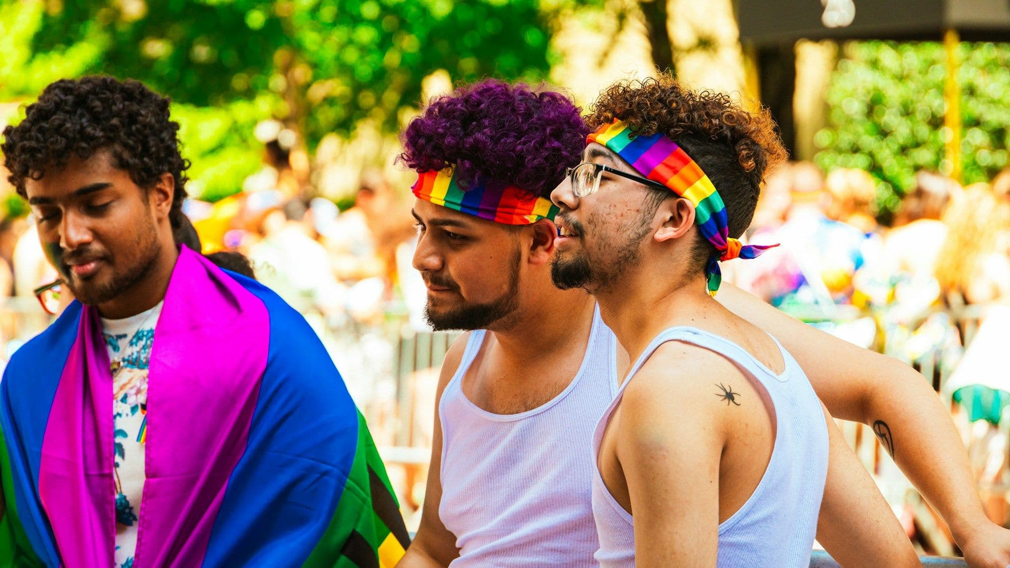 Three young men in white vests and rainbow headbands together at a Pride parade, one wrapped in a Pride flag.