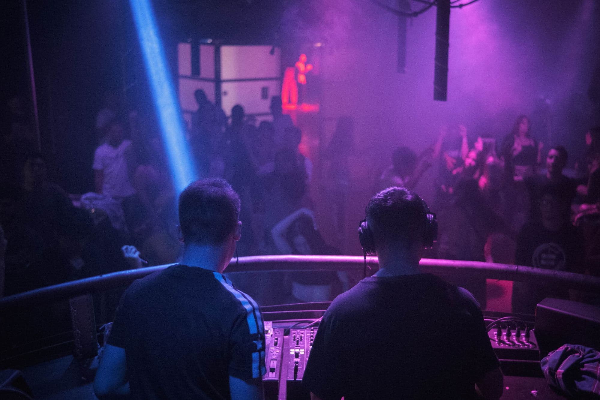 A DJ works above a packed dance floor under violet and blue lights.