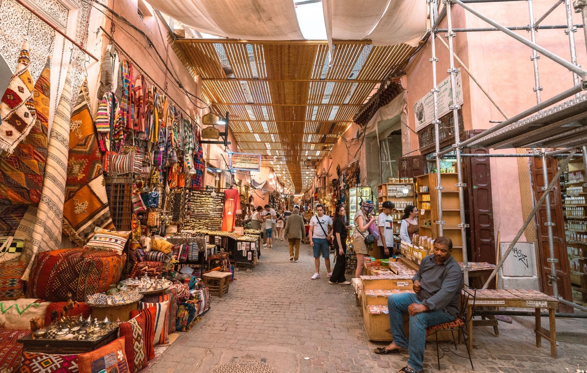 A covered souk lane in Marrakech lined with rugs, lanterns and textiles, with a shopkeeper seated on the right.
