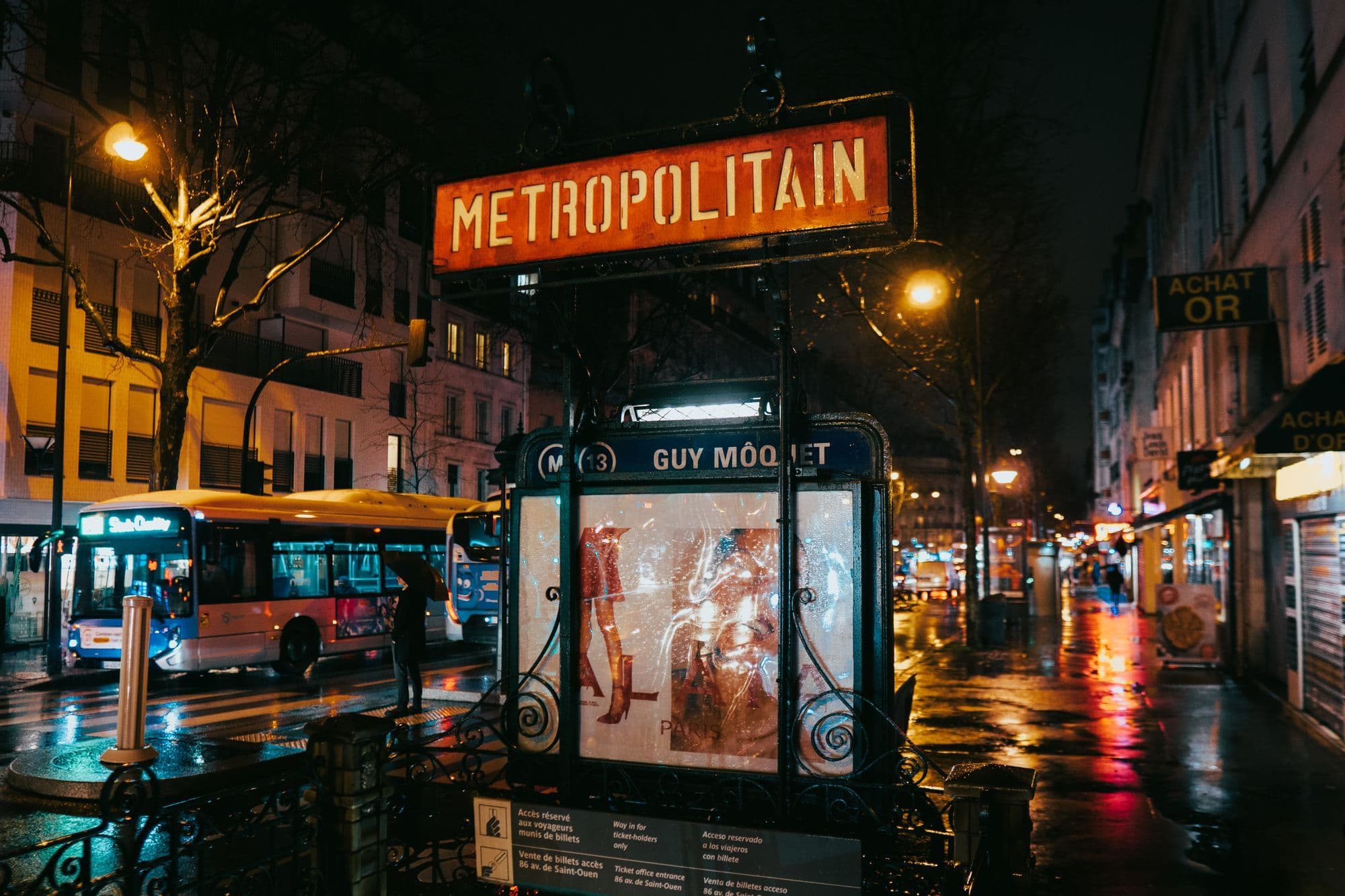 A rain-slick Paris street glows beside an Art Nouveau Metro entrance at night.
