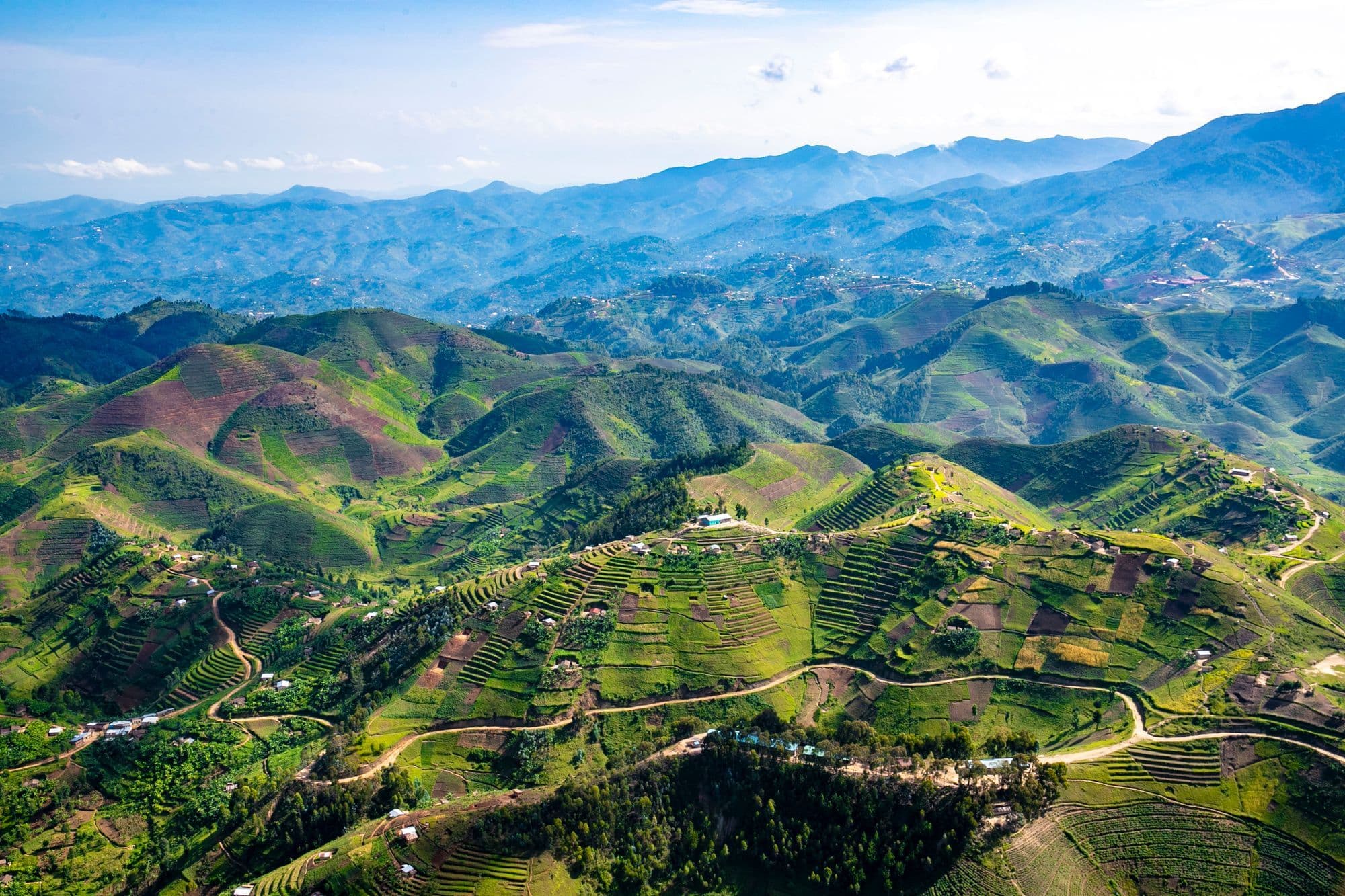 Aerial view of steep green hills covered in terraced fields and winding roads in the Rwandan countryside, with mountains beyond.