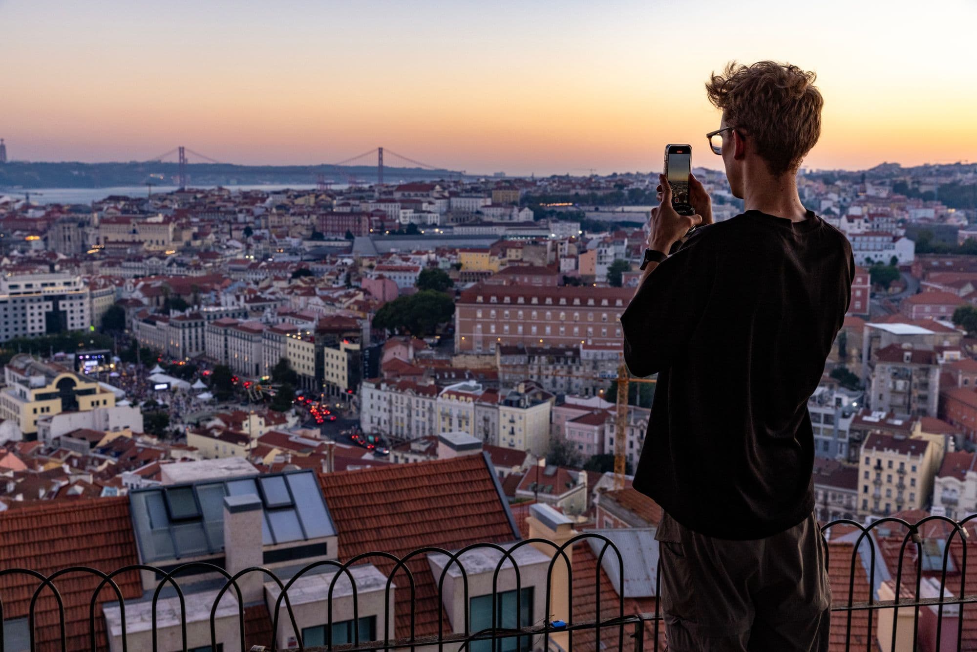 A man photographs the sunset from a viewpoint railing above Lisbon's rooftops, with a suspension bridge on the horizon.