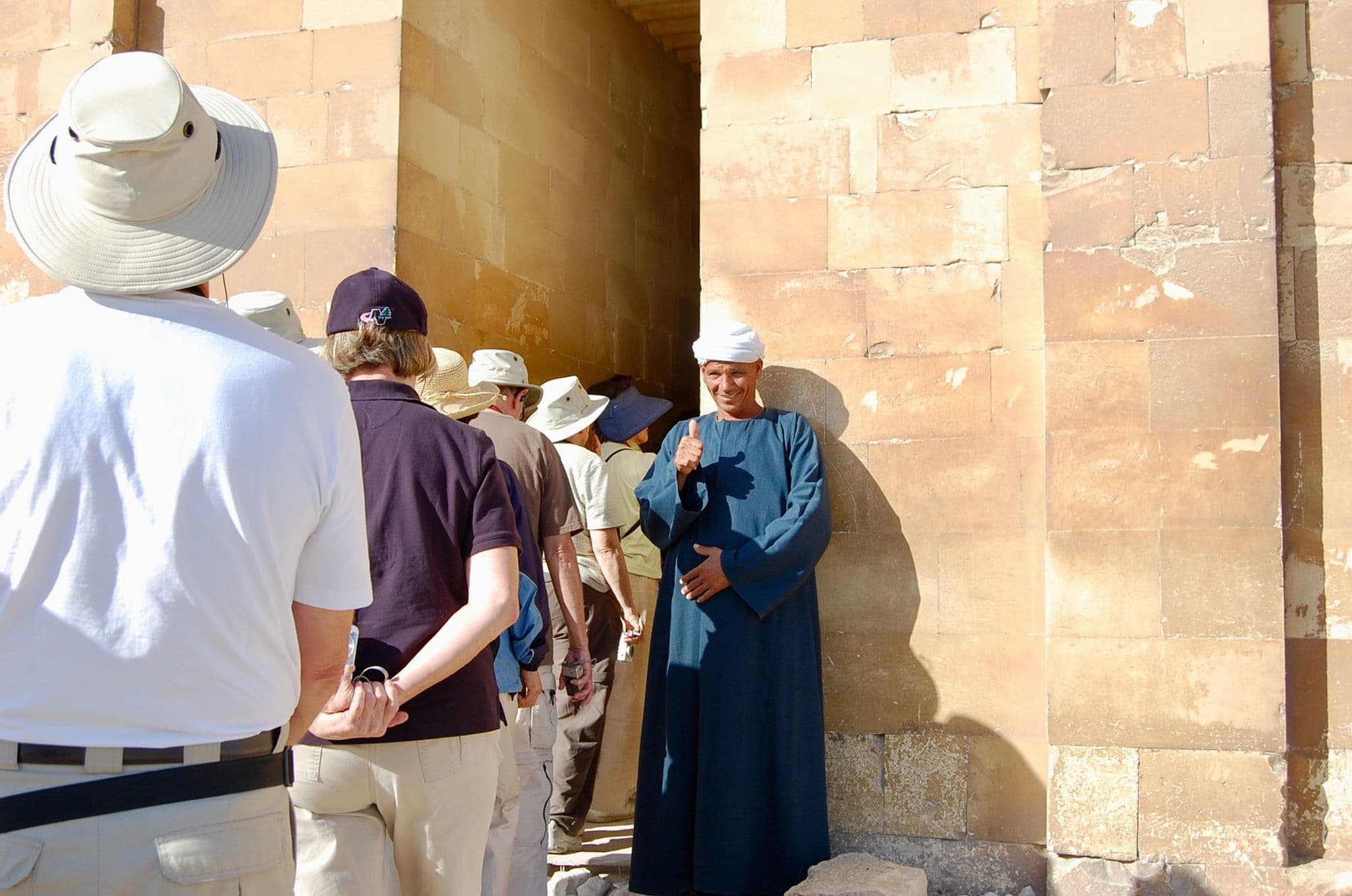 Tourists in sun hats pass a man in a pale blue galabiya standing at the stone doorway of an ancient Egyptian site.