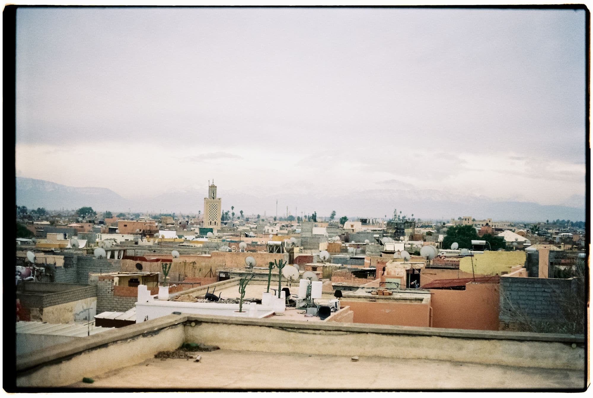 Flat rooftops and satellite dishes across the Marrakech medina, with the Koutoubia minaret and hazy mountains on the horizon.