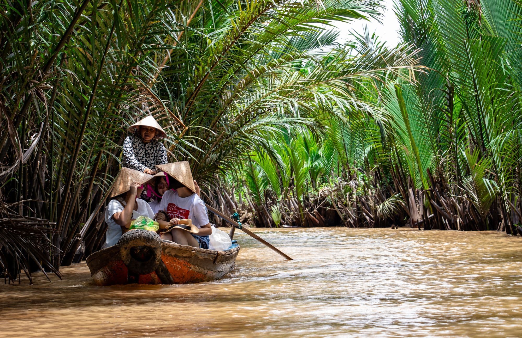 Two people in conical hats paddle a wooden boat along a narrow brown waterway lined with water palms.