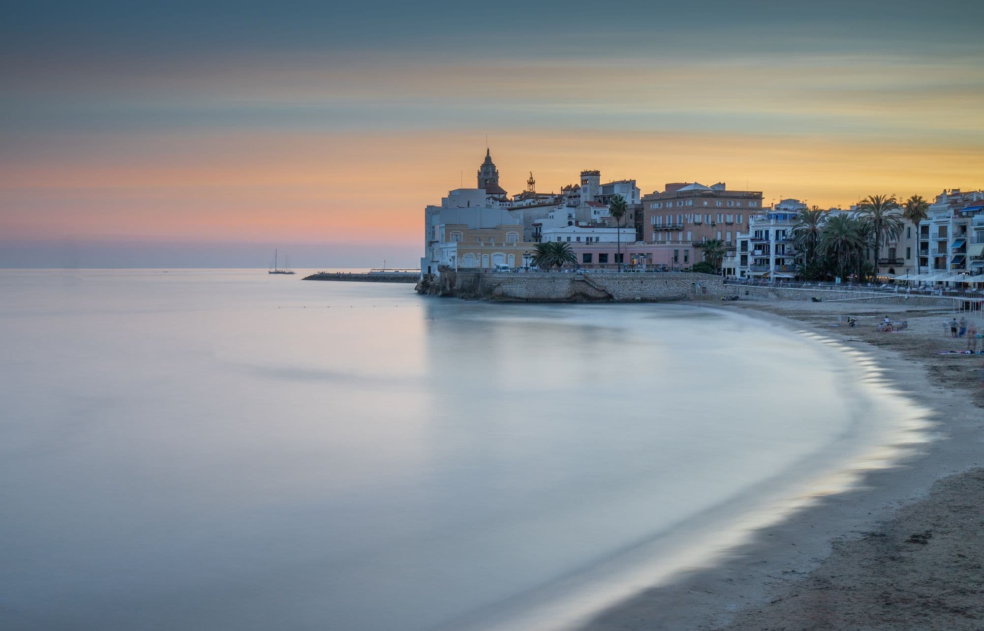 Sitges seafront at twilight, with the church on its headland above a curved sandy beach and calm sea.