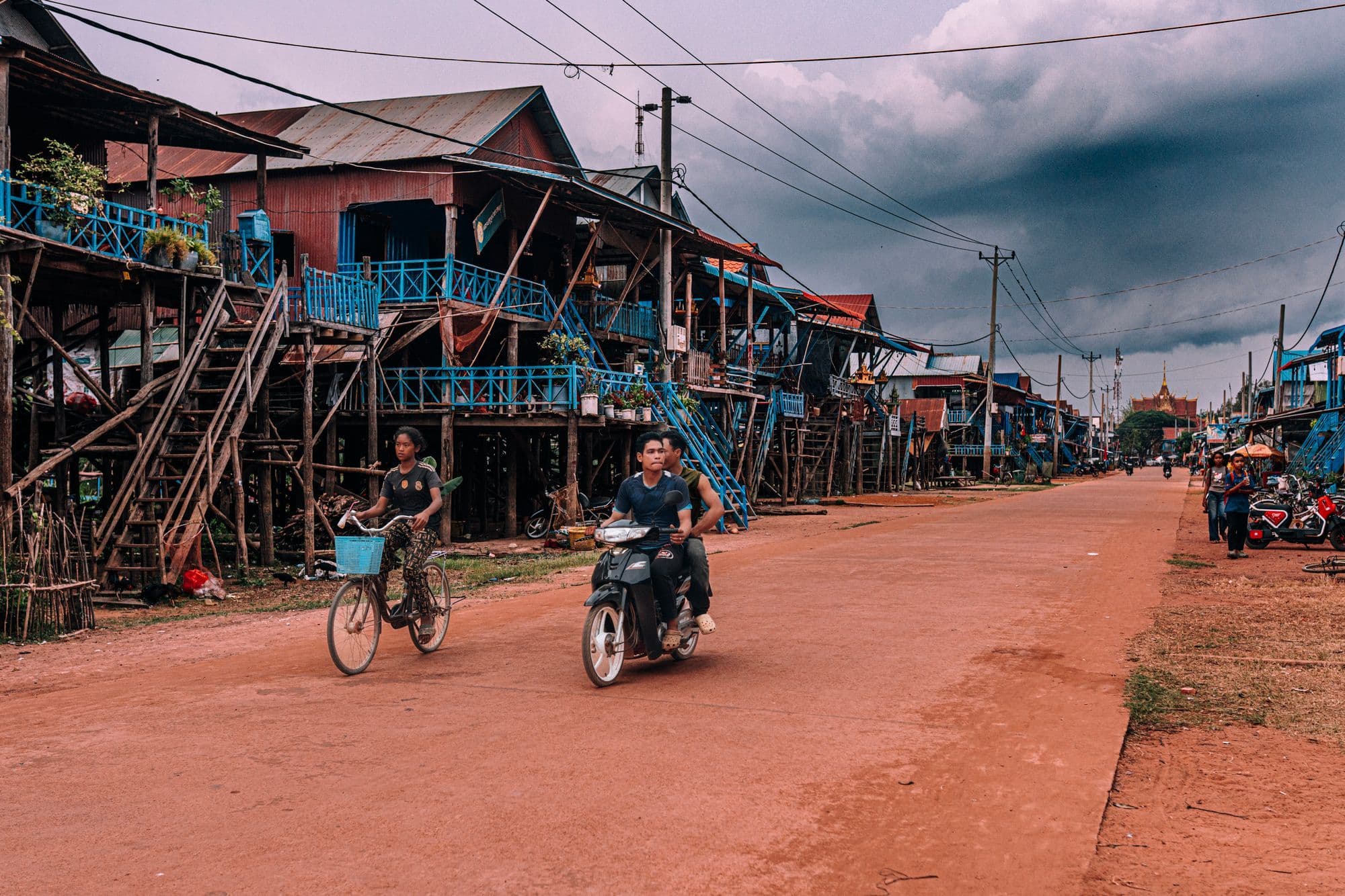 A cyclist and a motorbike rider on a red dirt road between tall wooden houses on stilts in Kampong Phluk.