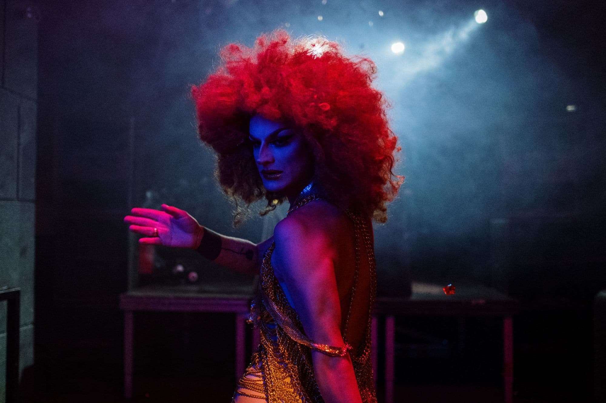 A drag performer poses on a lit stage in front of a nightclub audience.