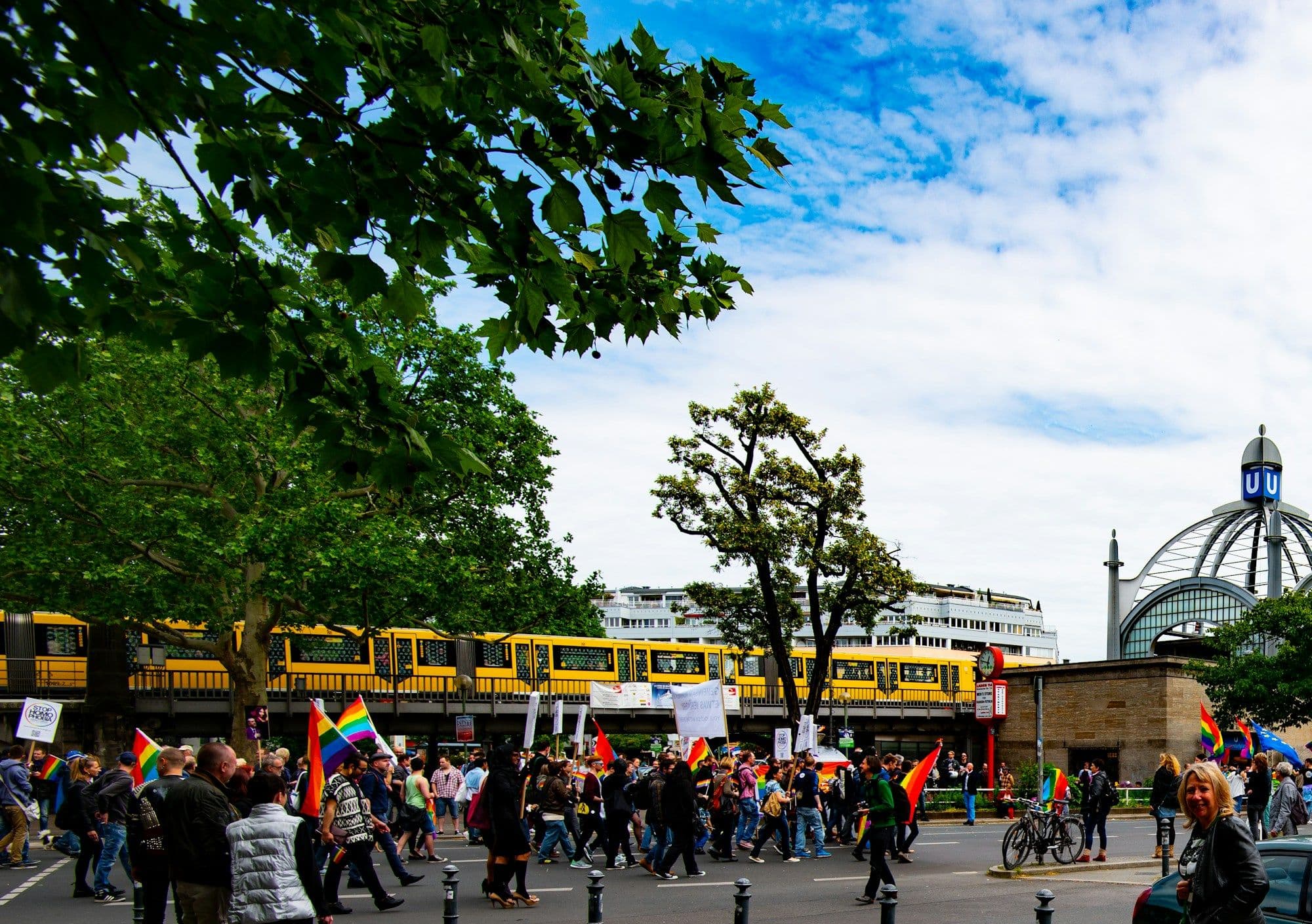 A Berlin Pride march moving along a tree-lined city street under rainbow flags.
