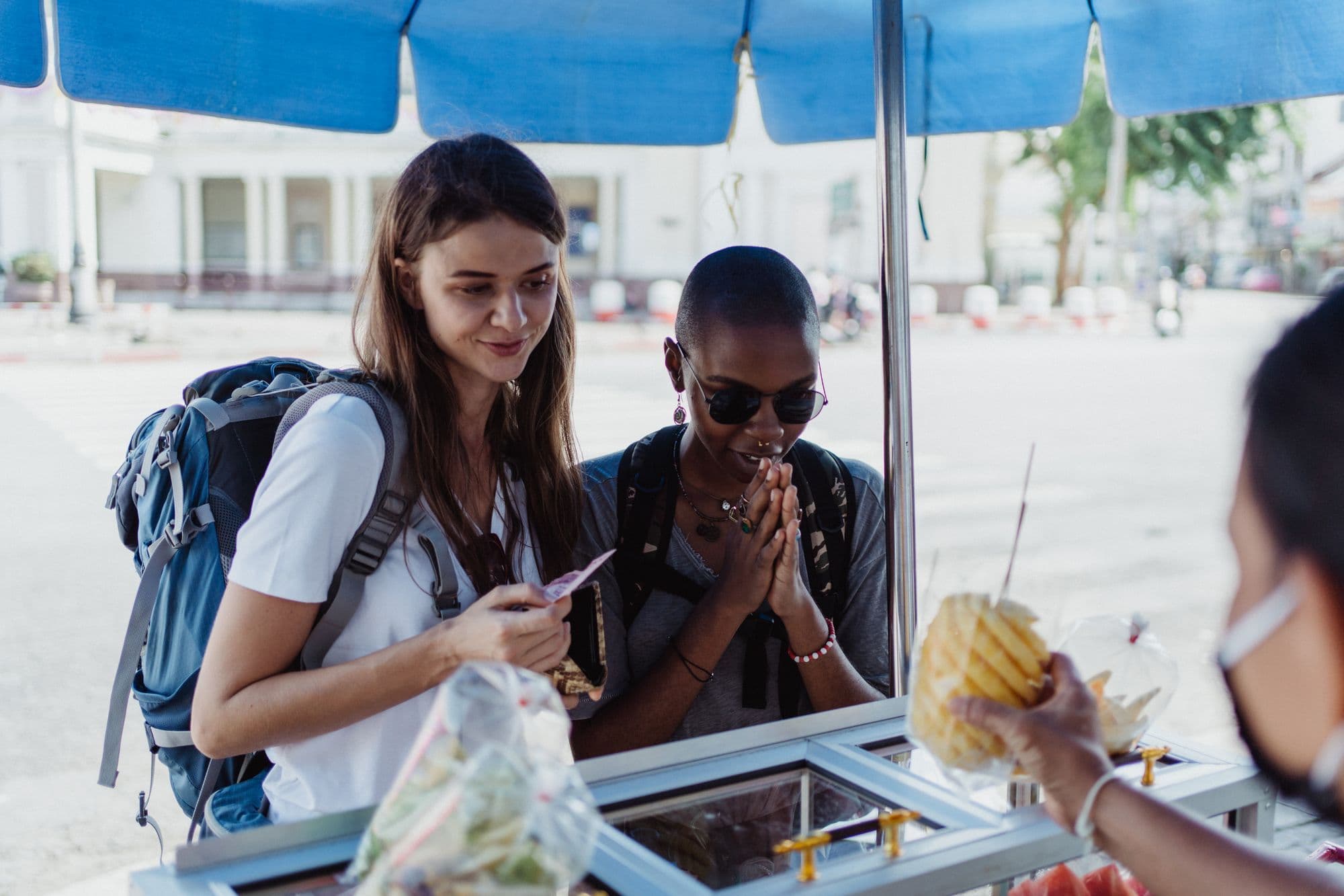 Two travelers buy food from a vendor at an outdoor stall.
