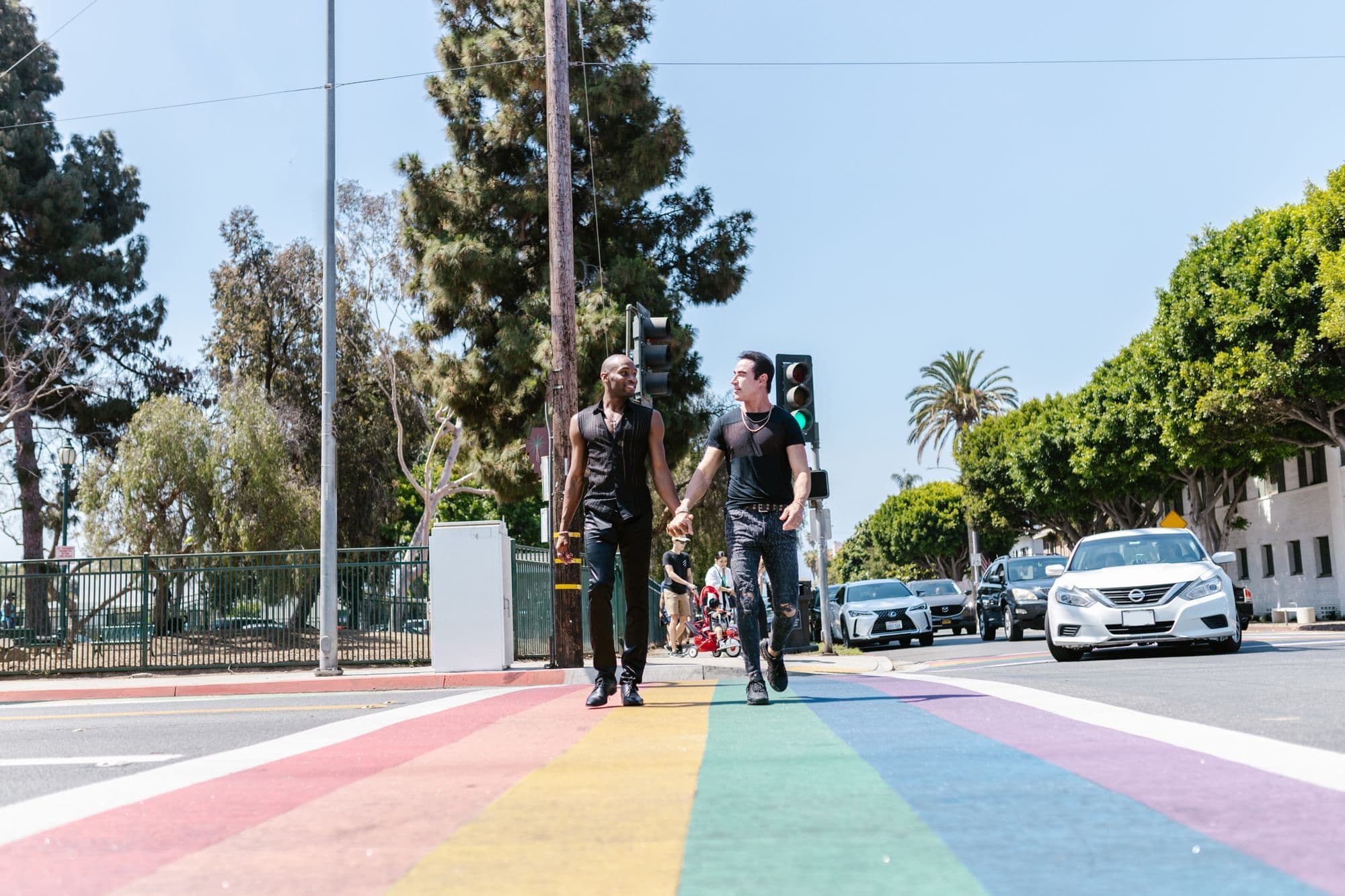 Two people hold hands while crossing a rainbow-painted city crosswalk.