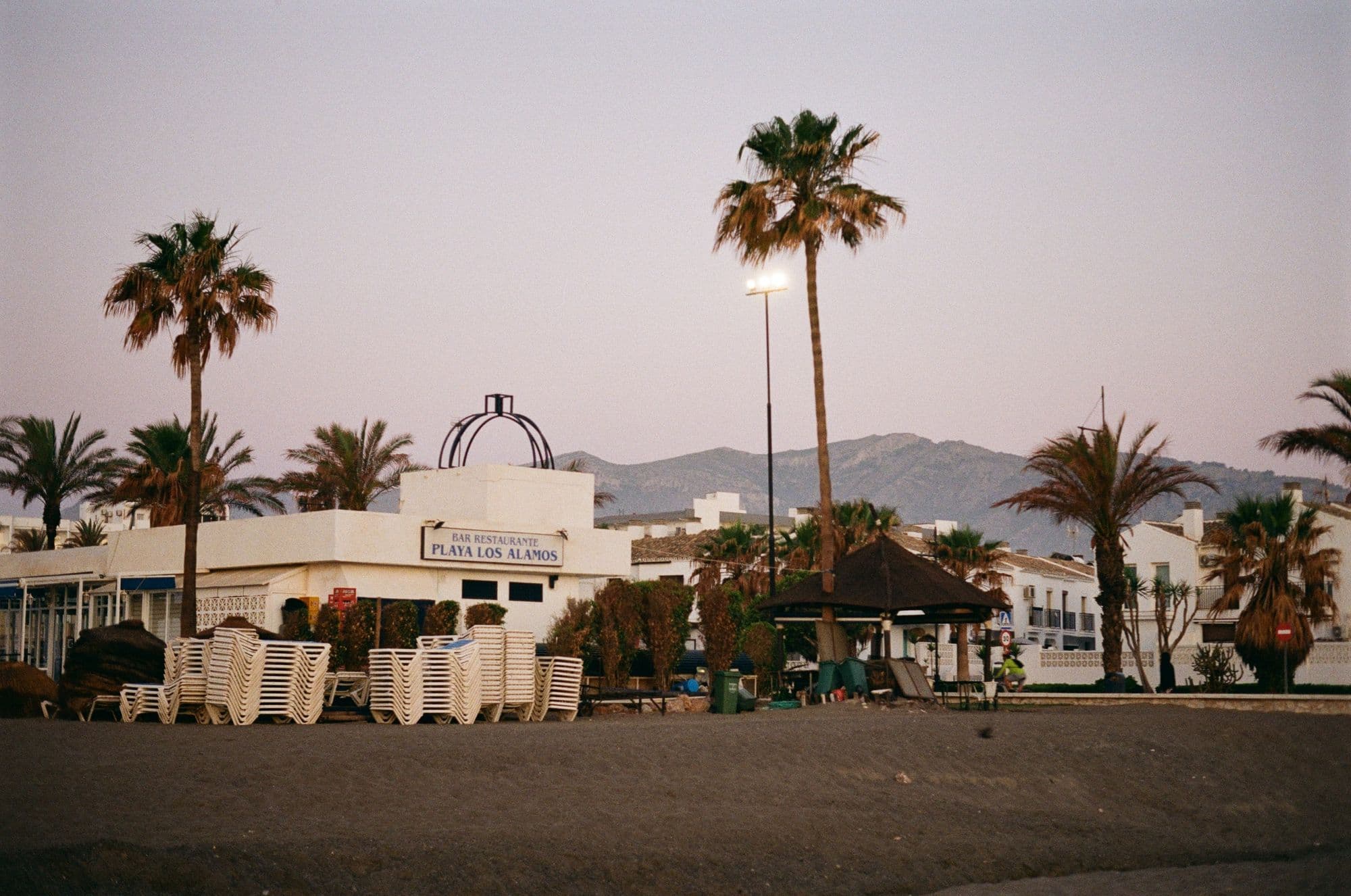 Palm trees and a white beach restaurant on Playa Los Álamos in Torremolinos at dusk, with mountains behind.