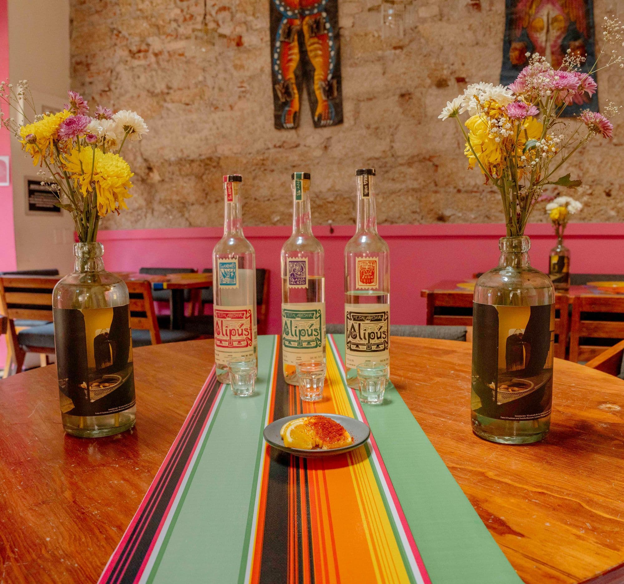 Three bottles of mezcal and small tasting glasses on a striped runner, with flowers in bottles and a pink wall behind.