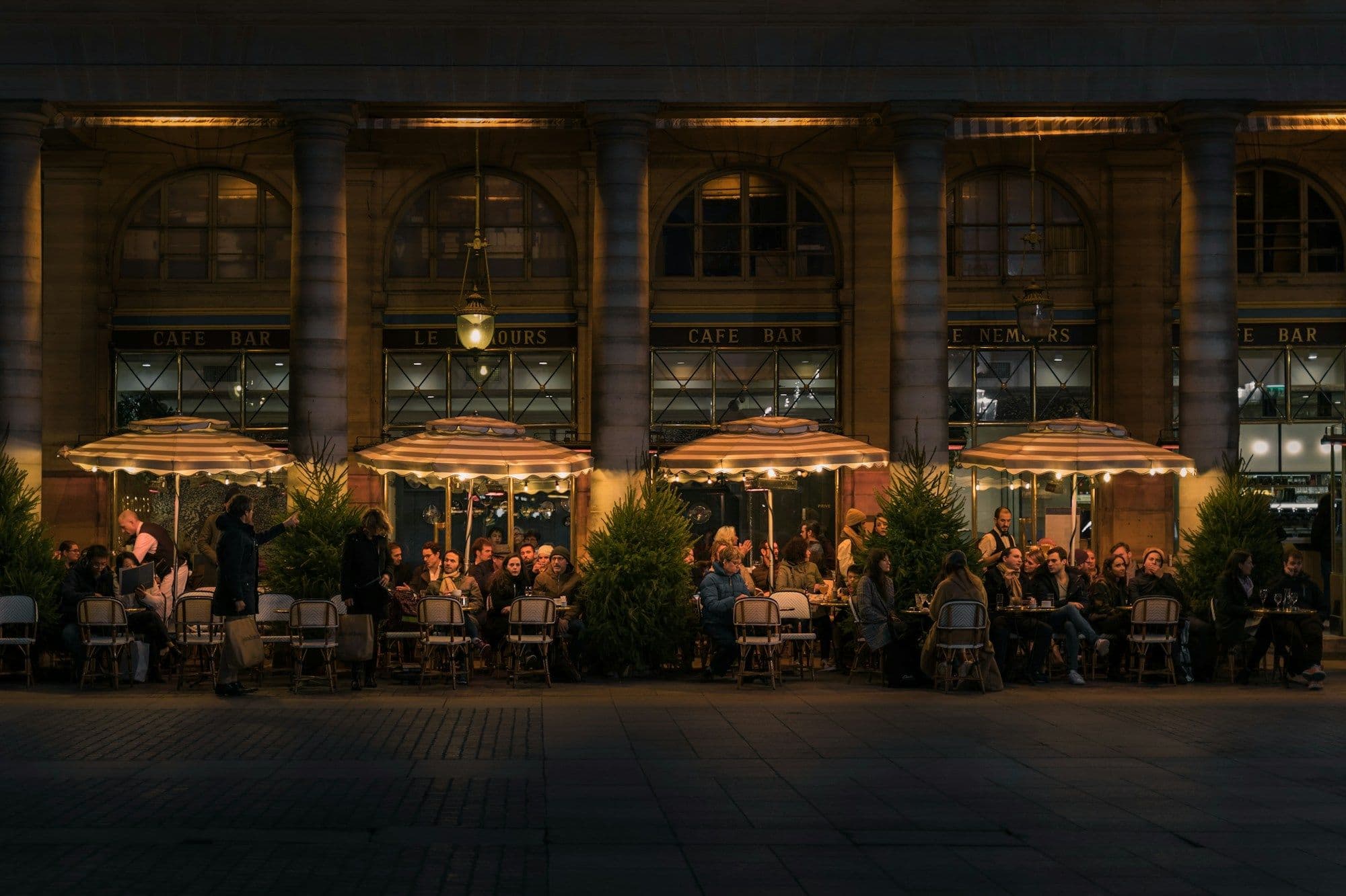 Diners sit at small tables beneath striped awnings outside a café after dark.