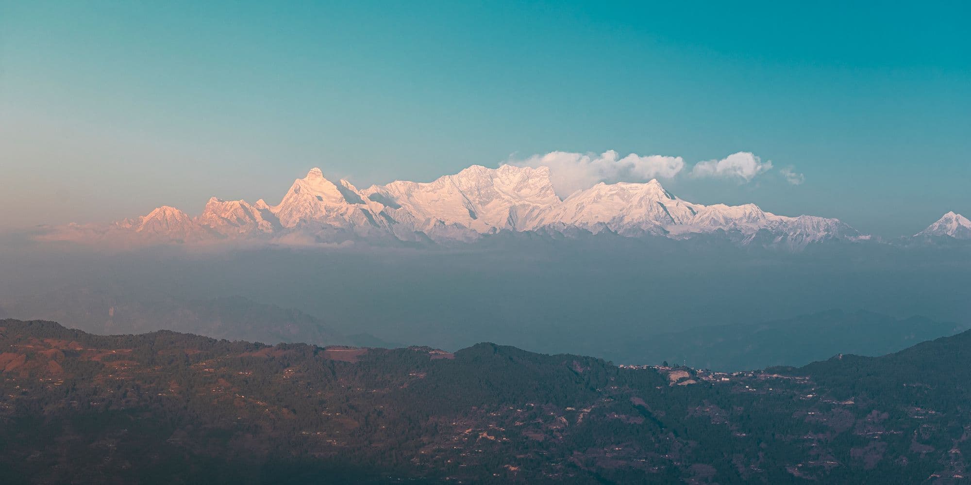 Snow-covered Himalayan peaks glowing pink above a hazy band of hills at dawn.