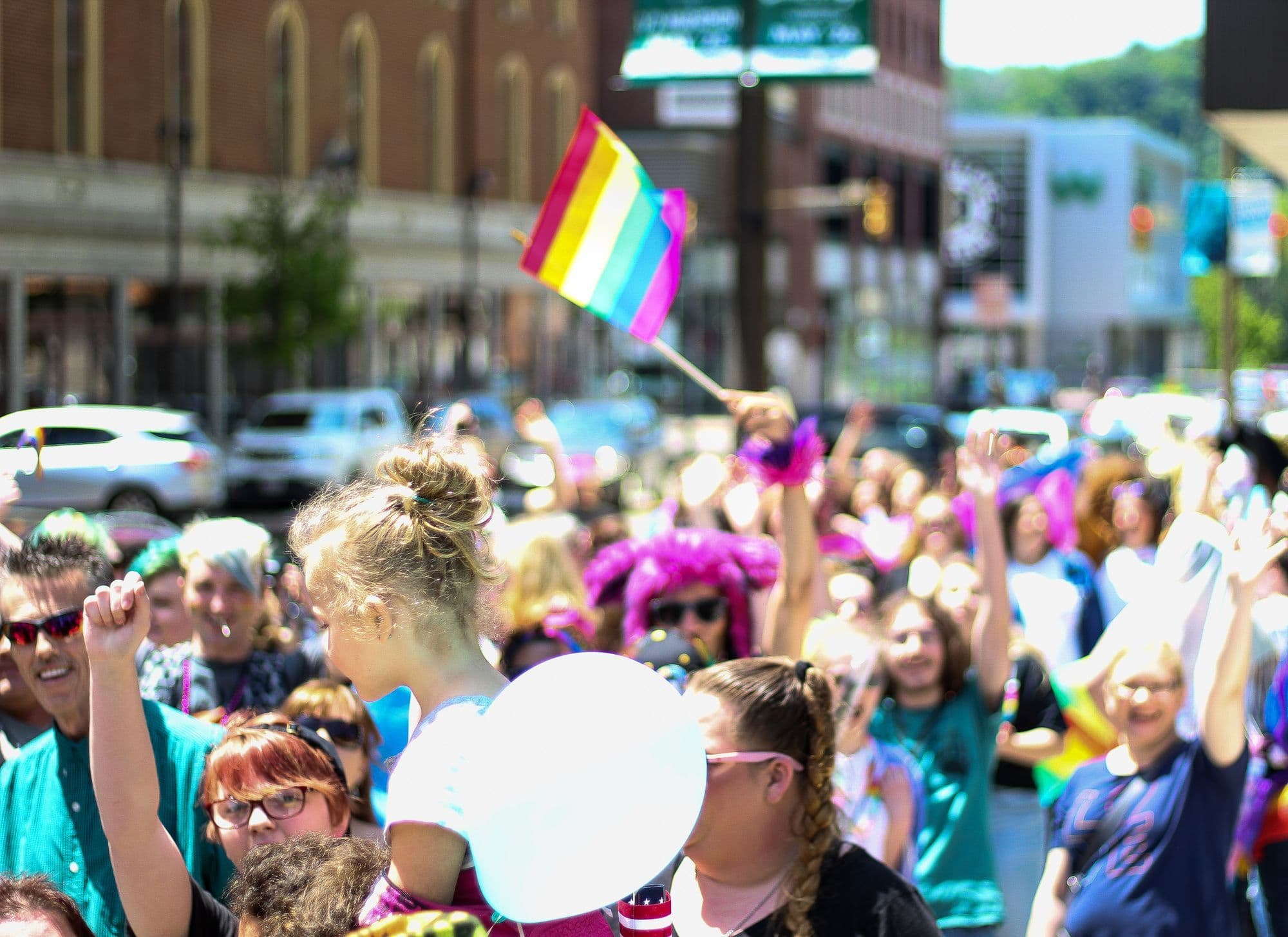 A large rainbow flag moves above a packed city street at a daytime Pride parade.