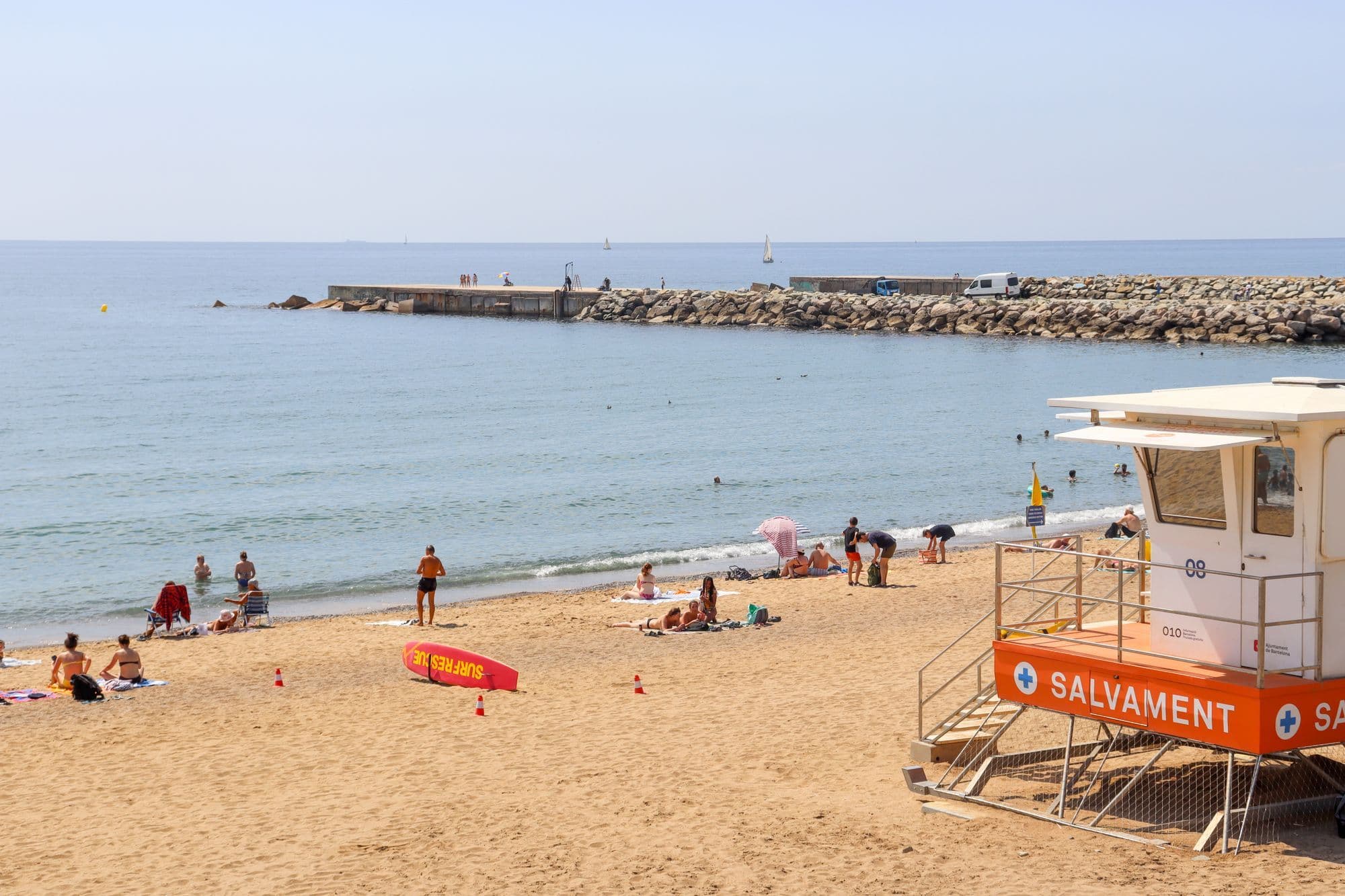 A sunny Barcelona city beach with sunbathers on the sand, an orange lifeguard tower and a stone breakwater in the sea.