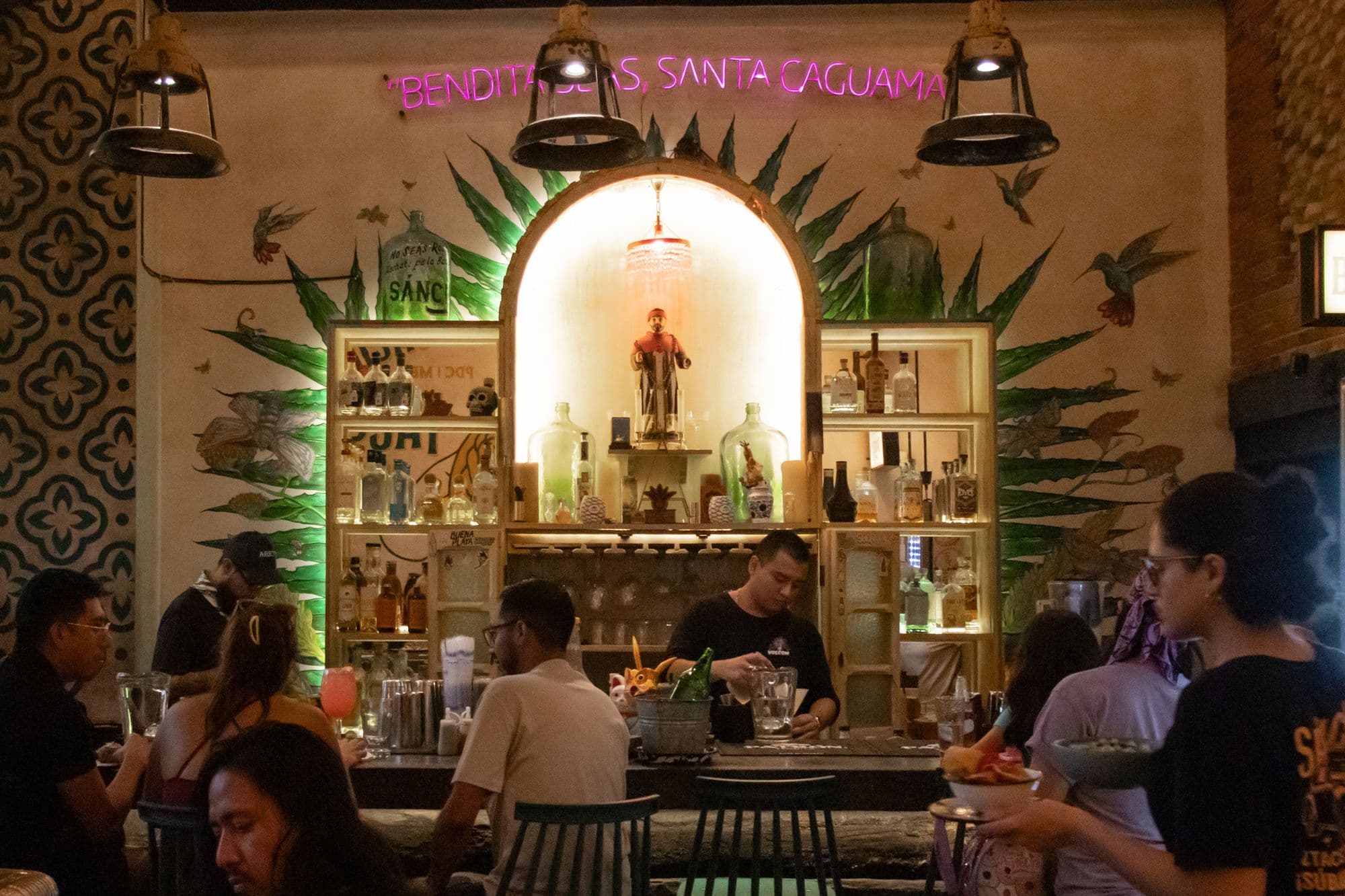 A busy Mexican bar at night, a bartender working in front of a lit arch of bottles and agave leaves painted on the wall.