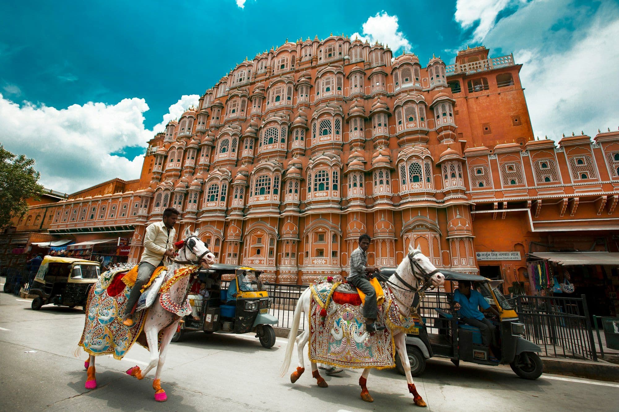 Two men in white ride decorated white horses past the pink sandstone facade of the Hawa Mahal in Jaipur, with auto-rickshaws behind.
