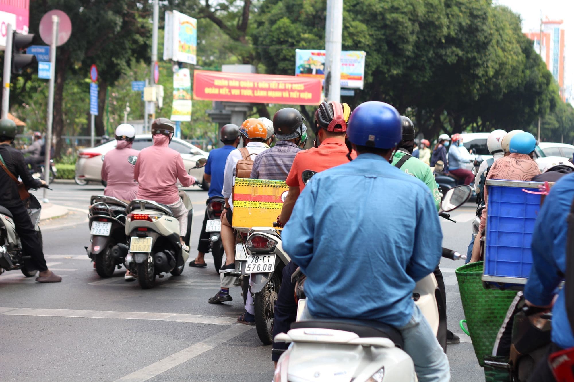 Riders in helmets wait on motorbikes at a busy Ho Chi Minh City junction on a sunny morning.