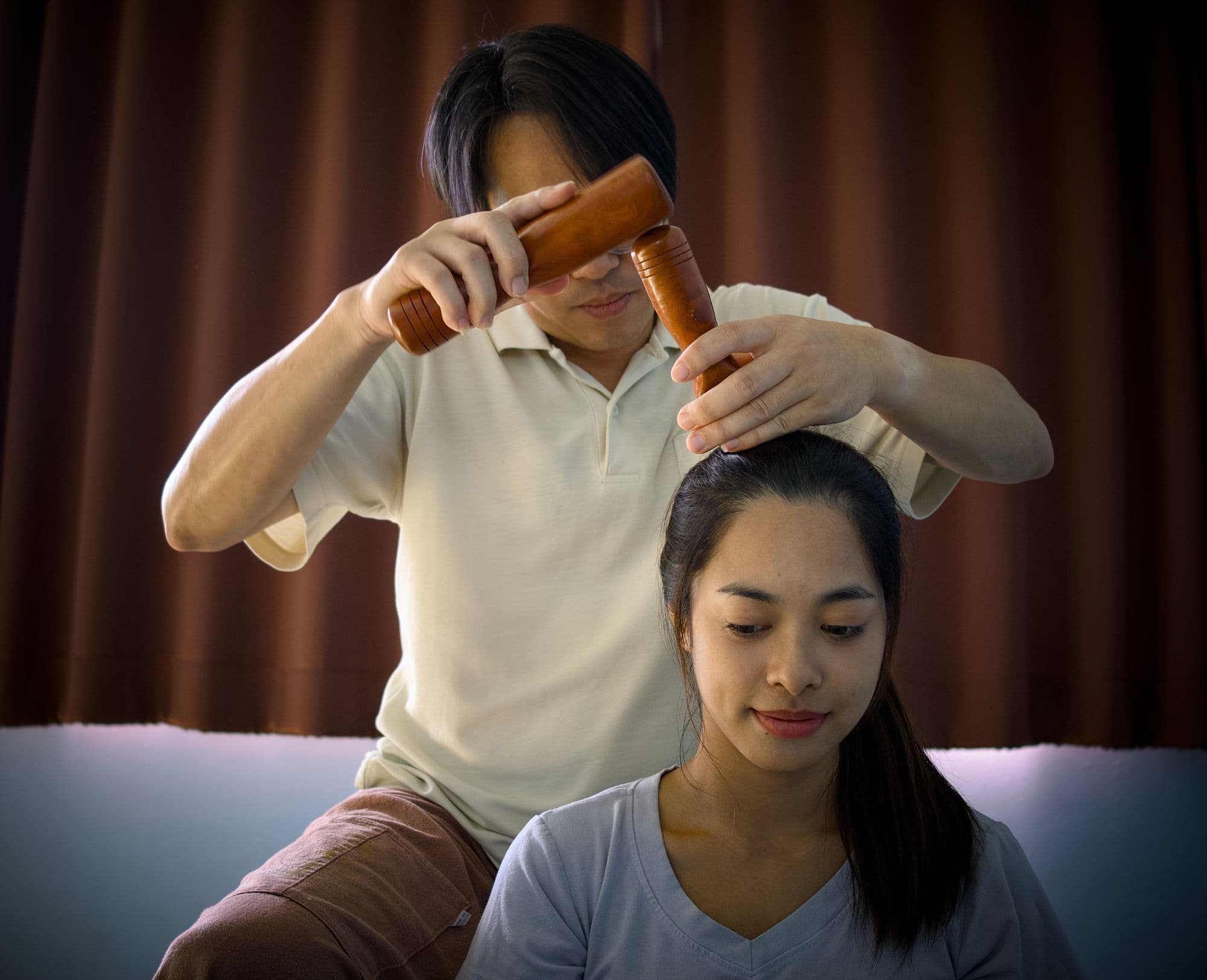 A Thai massage therapist working on a seated client's head with a traditional wooden tok sen tool.