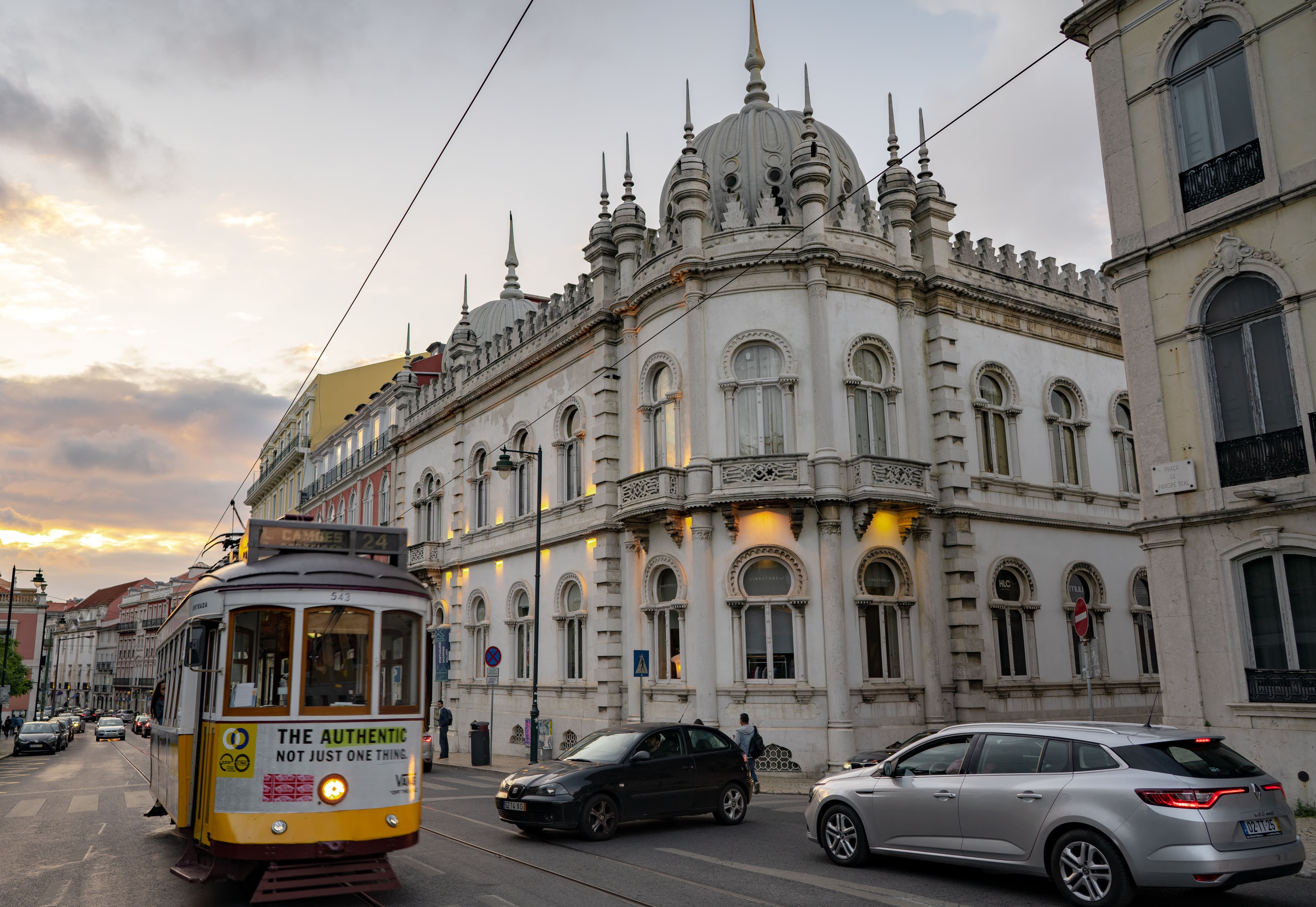 A yellow Lisbon tram passes the white Moorish-revival palace beside the Jardim do Príncipe Real at dusk.