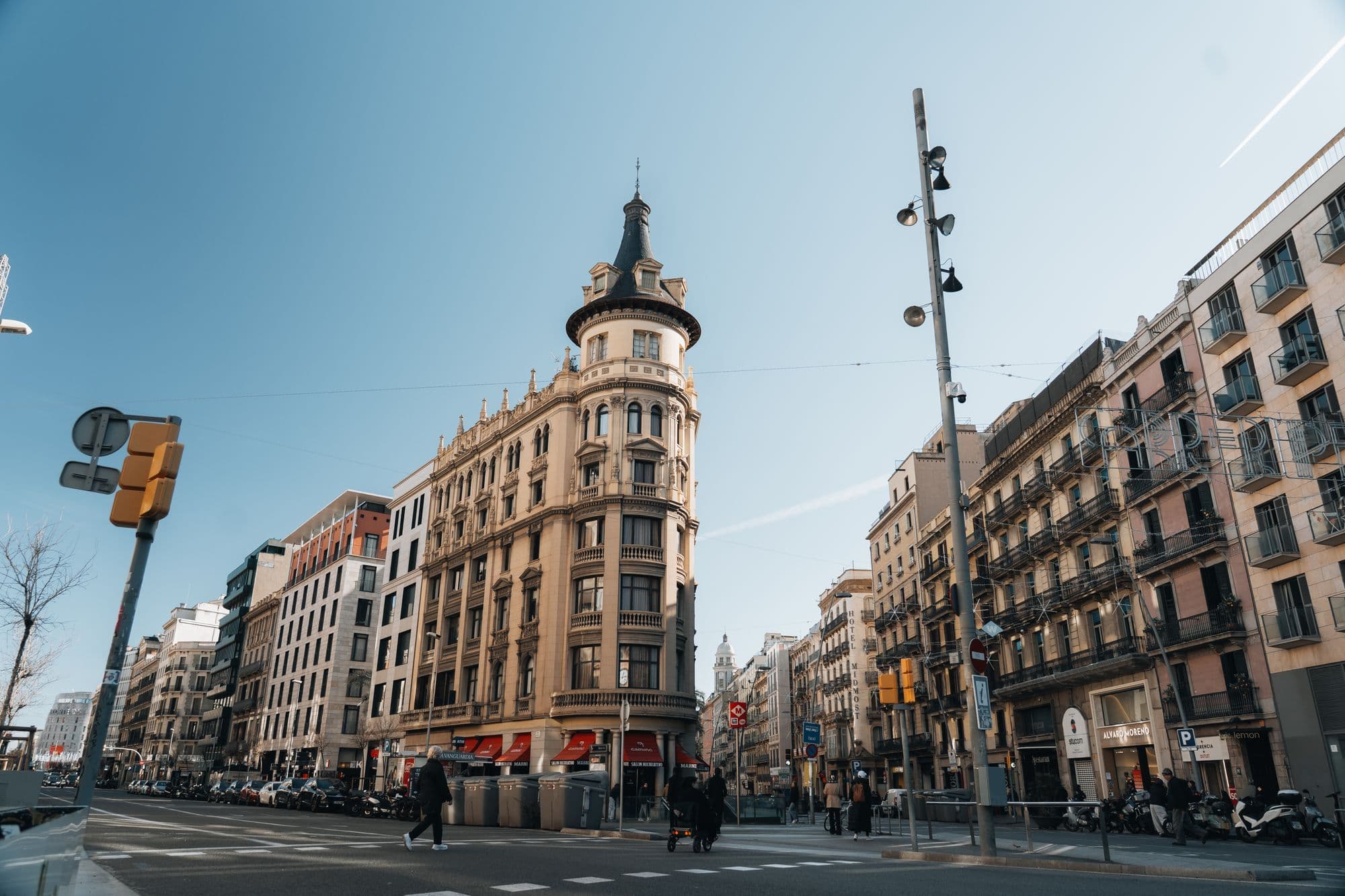 A historic Barcelona building stands at a sunny intersection beneath balconies and shutters.
