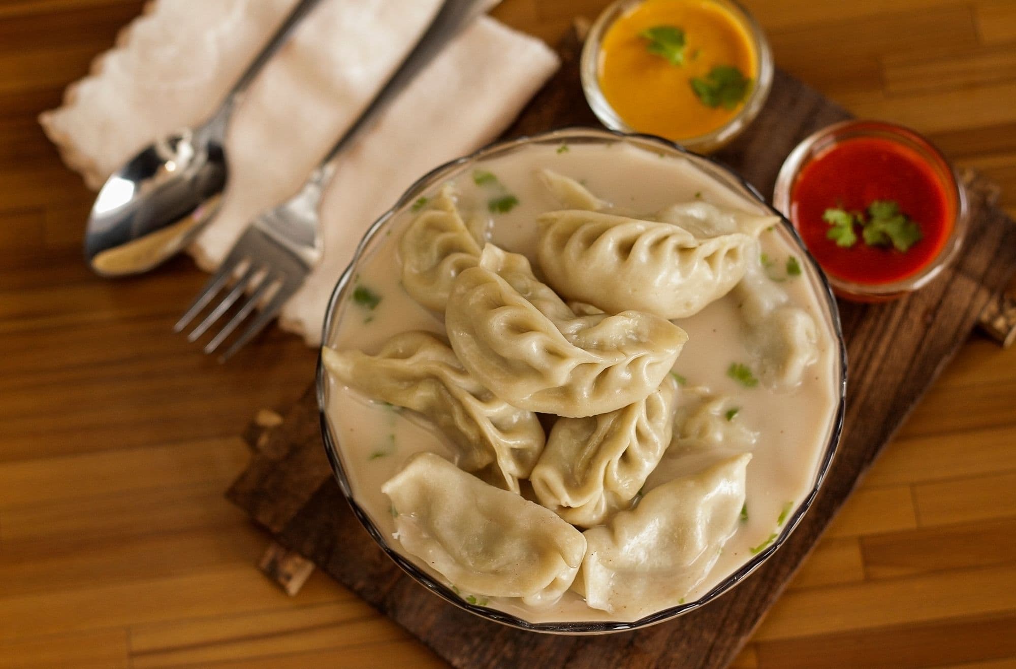 A bowl of pleated momo dumplings in sauce on a wooden board, with small bowls of chutney beside it.