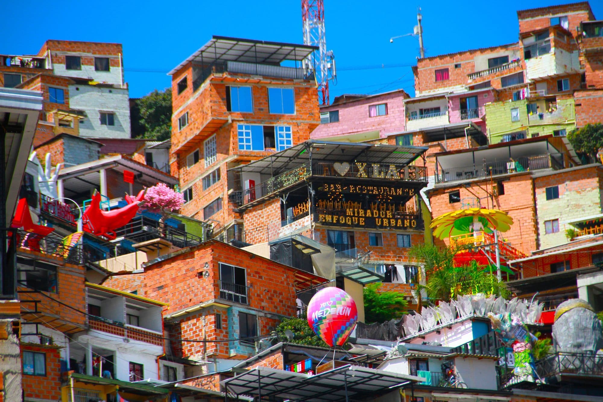 Brightly painted houses, murals and decorations stacked up a hillside in Comuna 13, Medellín, under a blue sky.