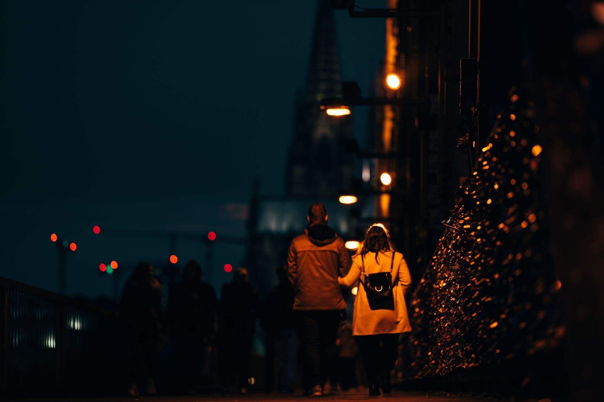 A couple walks along an illuminated Cologne street at night with the cathedral in the distance.