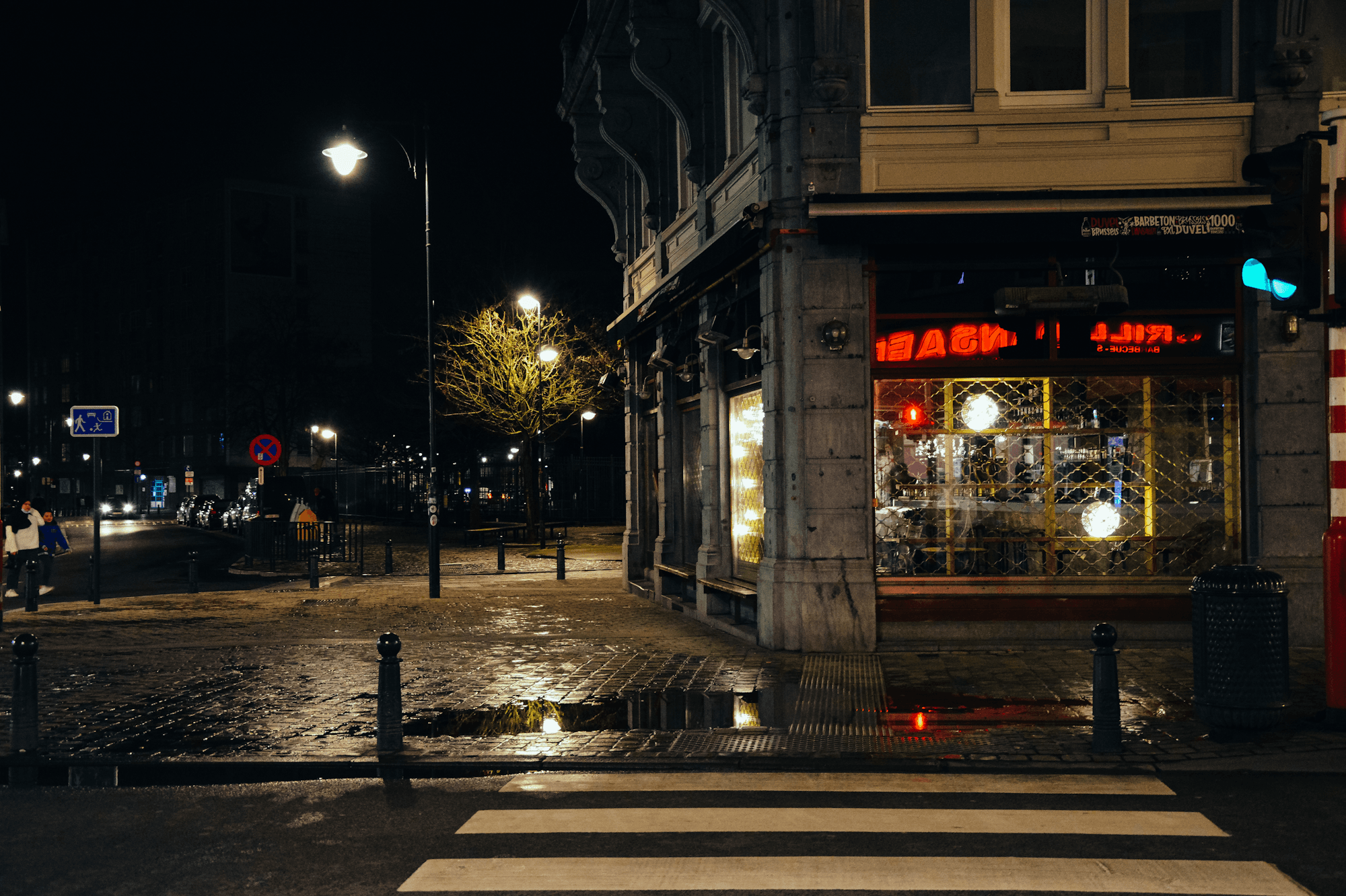 Pedestrians walking past lit shopfronts on a city street at night.