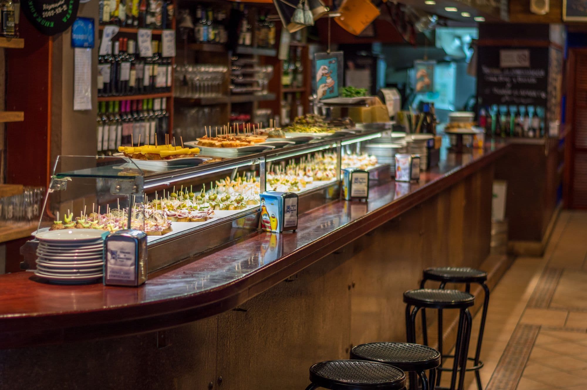 A long wooden tapas bar counter in Barcelona lined with plates of pintxos under glass, with empty stools in front.