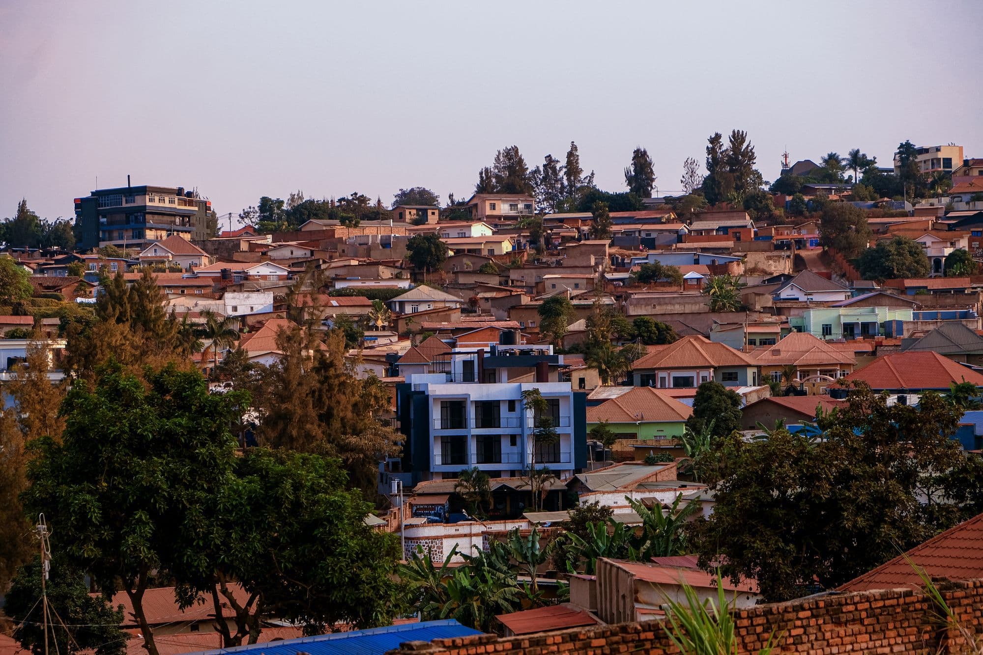 Red-roofed houses and new buildings climbing a green hillside in Kigali in warm evening light.