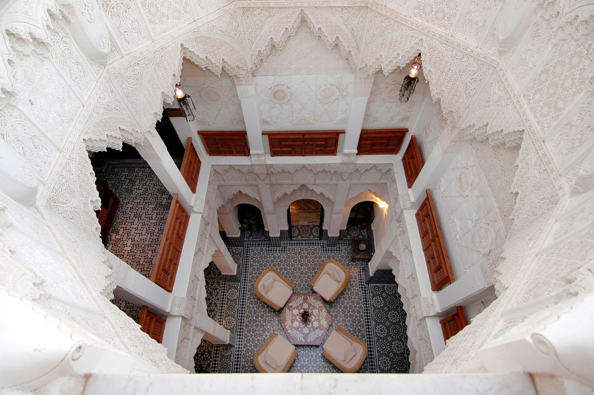 Looking down into the courtyard of a Marrakech riad, with carved white plaster balconies around a tiled floor and armchairs.