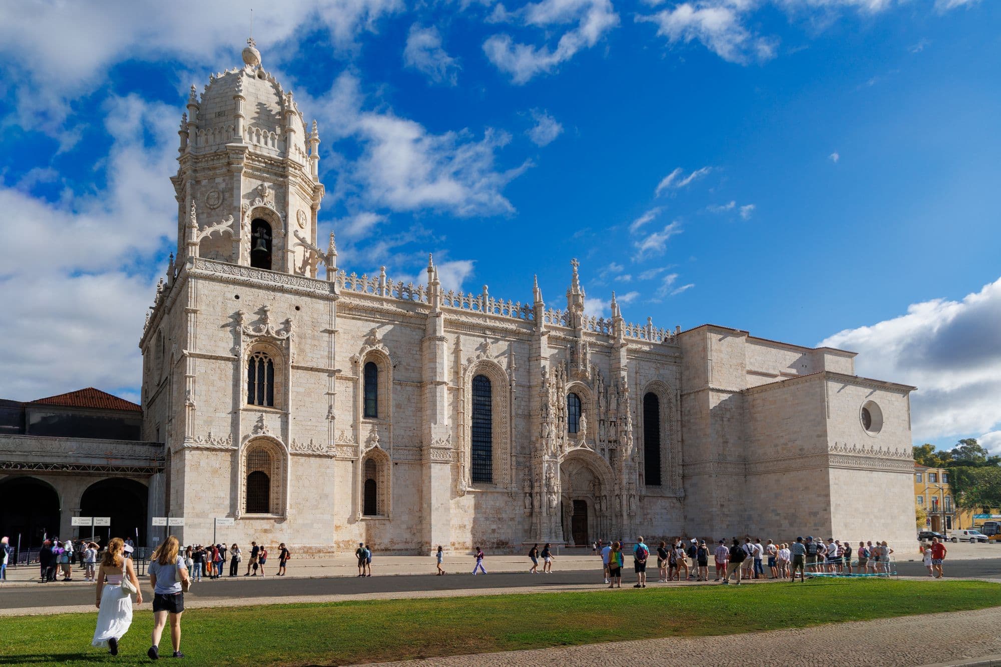 Visitors walk past the long carved limestone facade and bell tower of the Jerónimos Monastery under a blue sky.