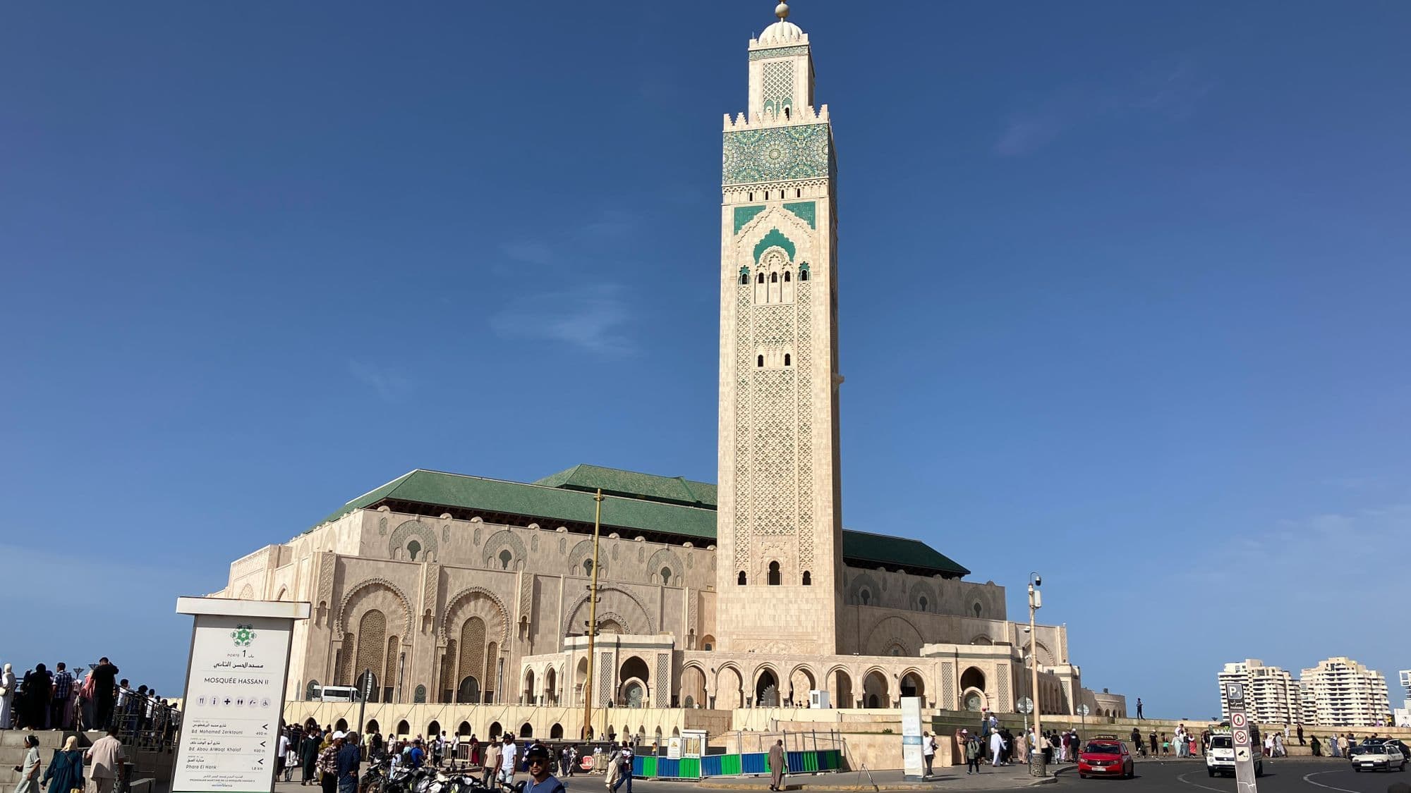 The tiled minaret and green-roofed prayer hall of the Hassan II Mosque under a clear blue sky, with visitors on the esplanade.