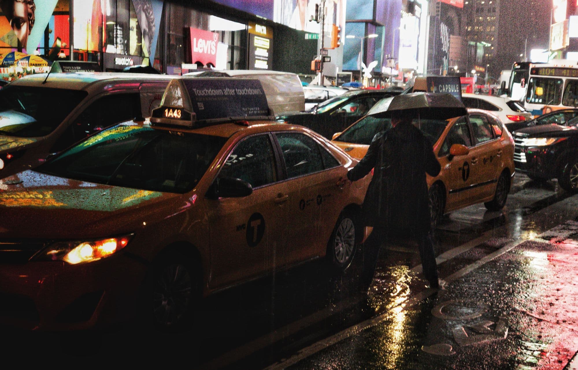 Yellow taxis pass through a wet city street under neon signs at night.
