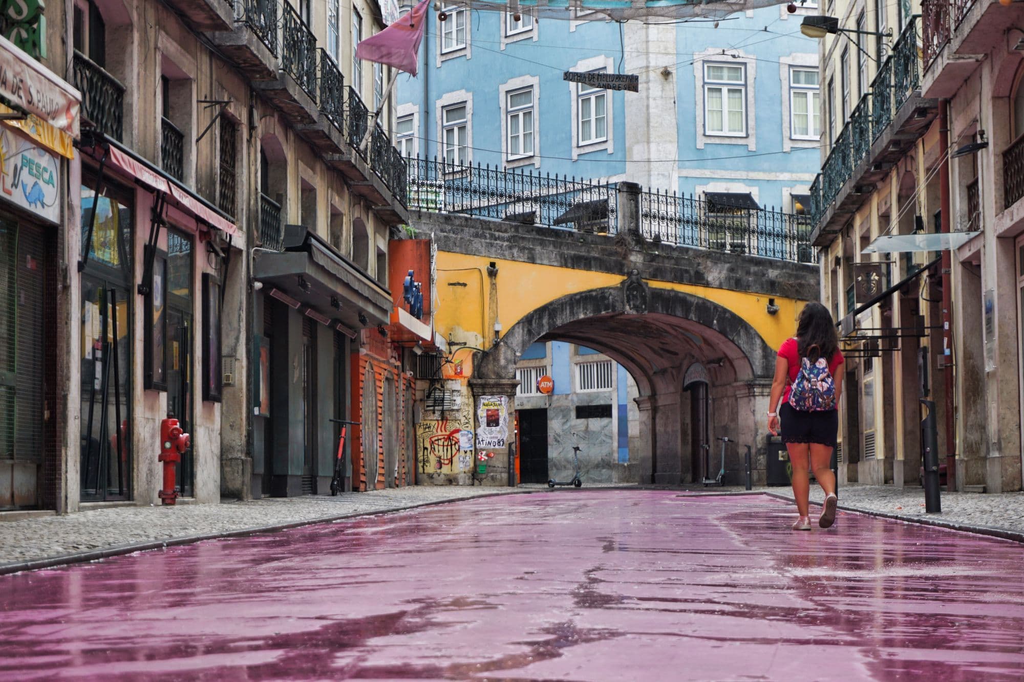 A woman with a backpack walks down Rua Nova do Carvalho, Lisbon's pink-painted street, under a yellow stone arch.