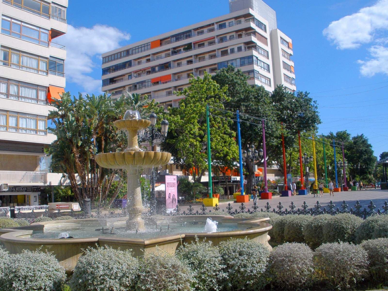 A fountain in the middle of Plaza de la Nogalera in Torremolinos, surrounded by palms, colourful poles and white apartment blocks.