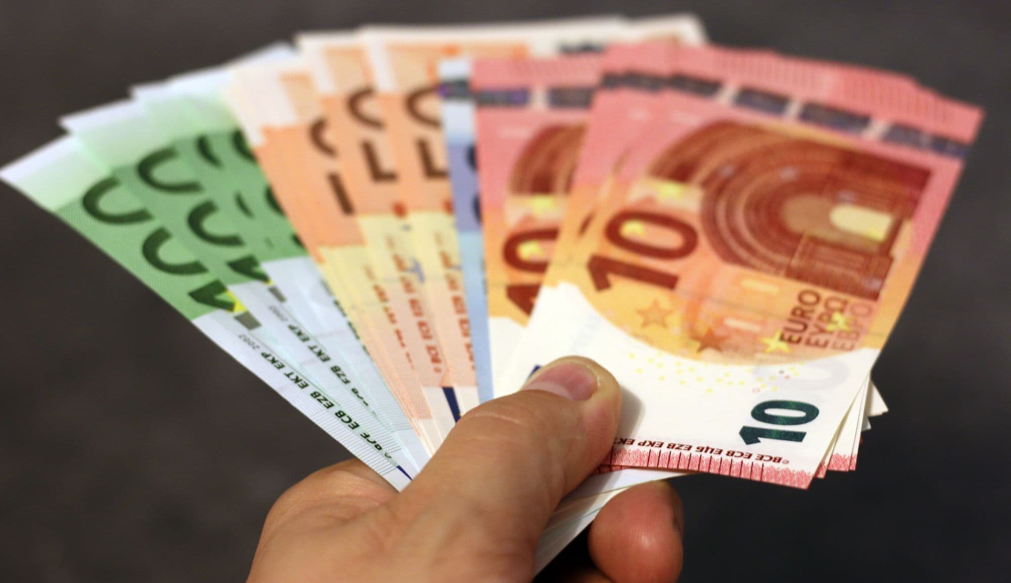 A hand holds a fan of euro banknotes in several colours against a dark background.