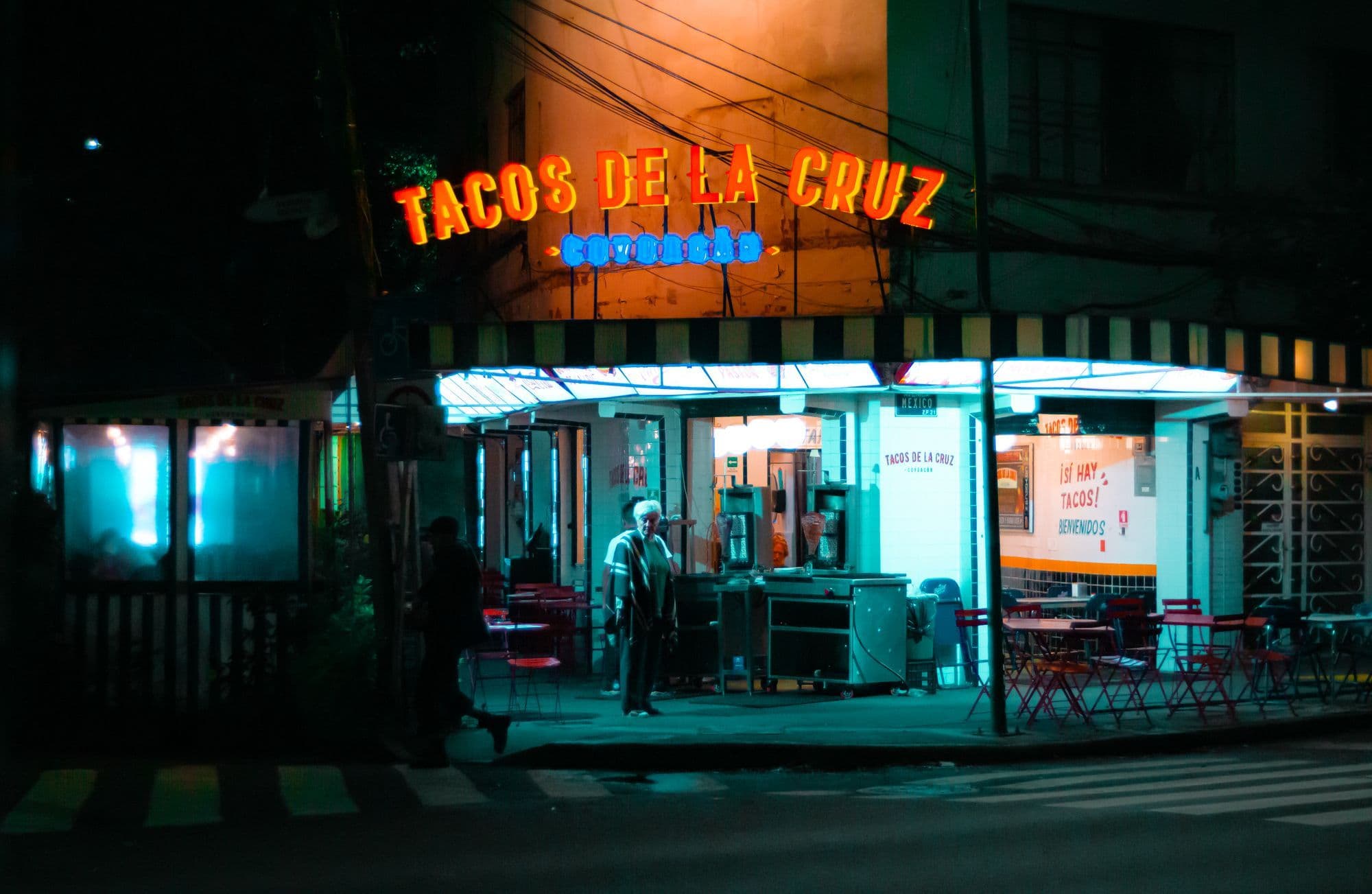 A corner taquería lit by an orange neon sign at night, a man standing by the counter and red tables on the pavement.
