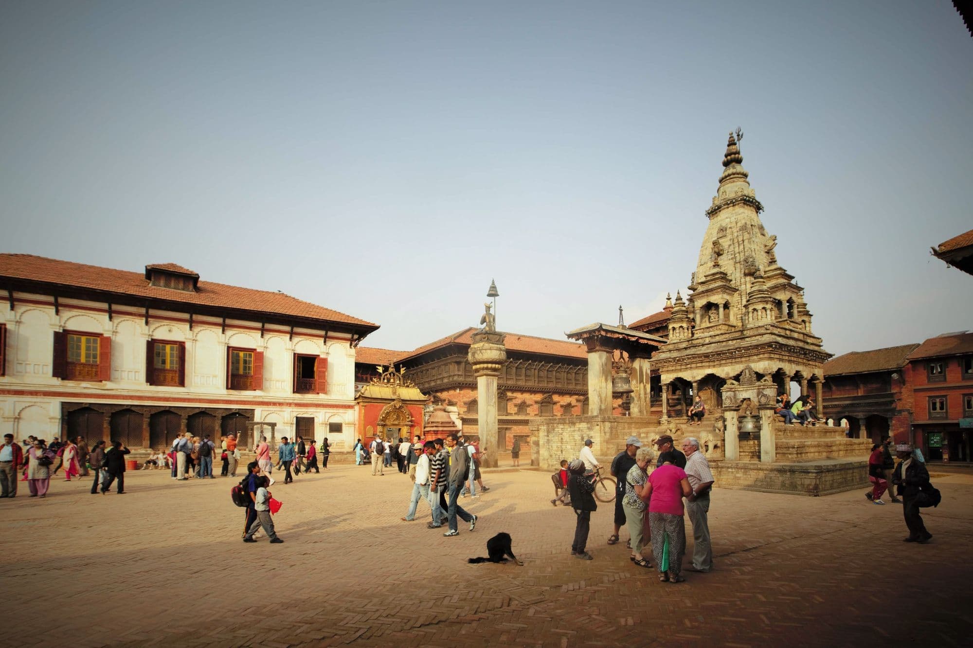 Visitors walk across Bhaktapur Durbar Square past brick palace buildings and a tall tiered stone temple.