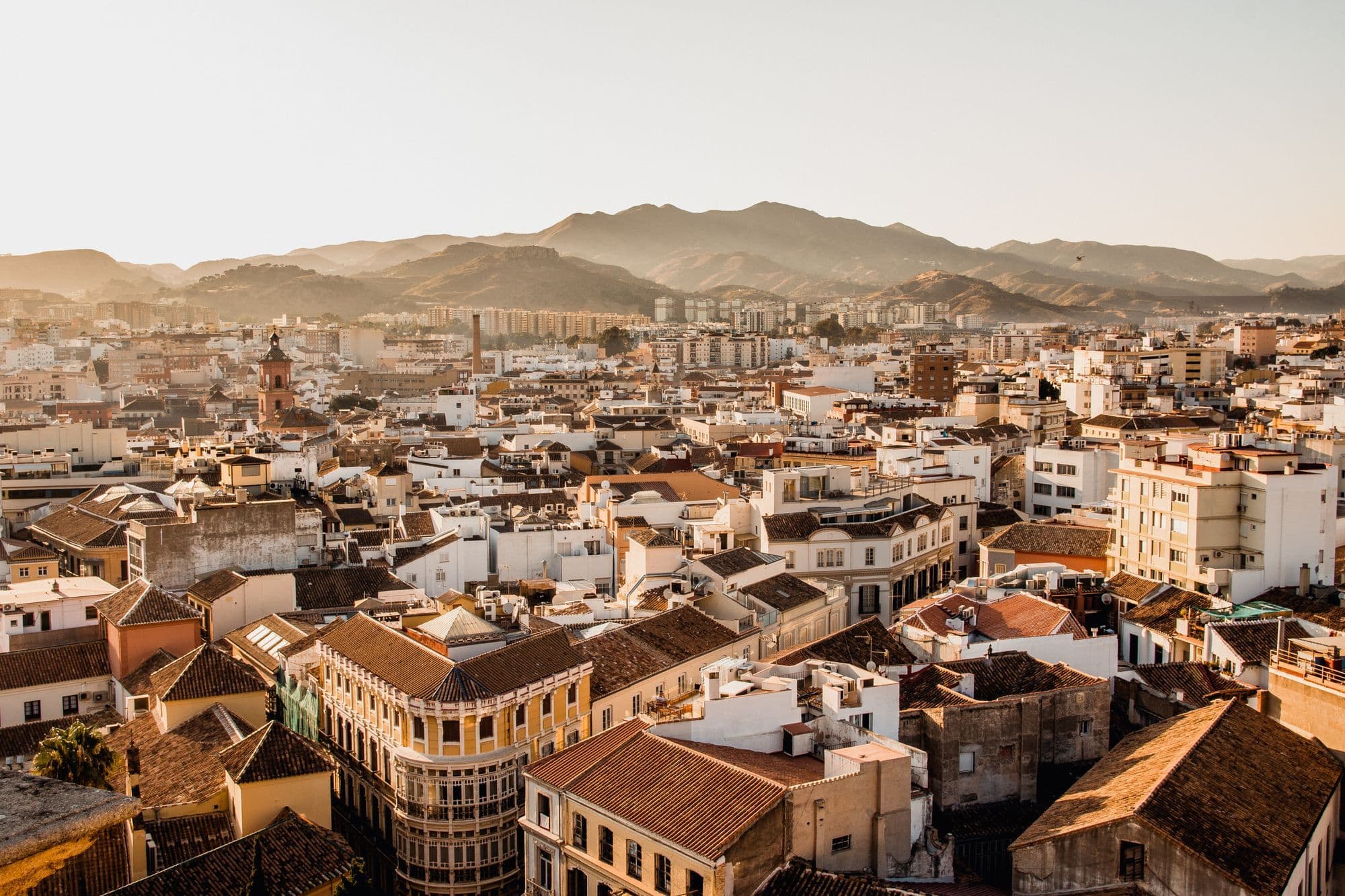 Rooftops of central Málaga at sunset, with a domed church tower and the mountains rising behind the city.