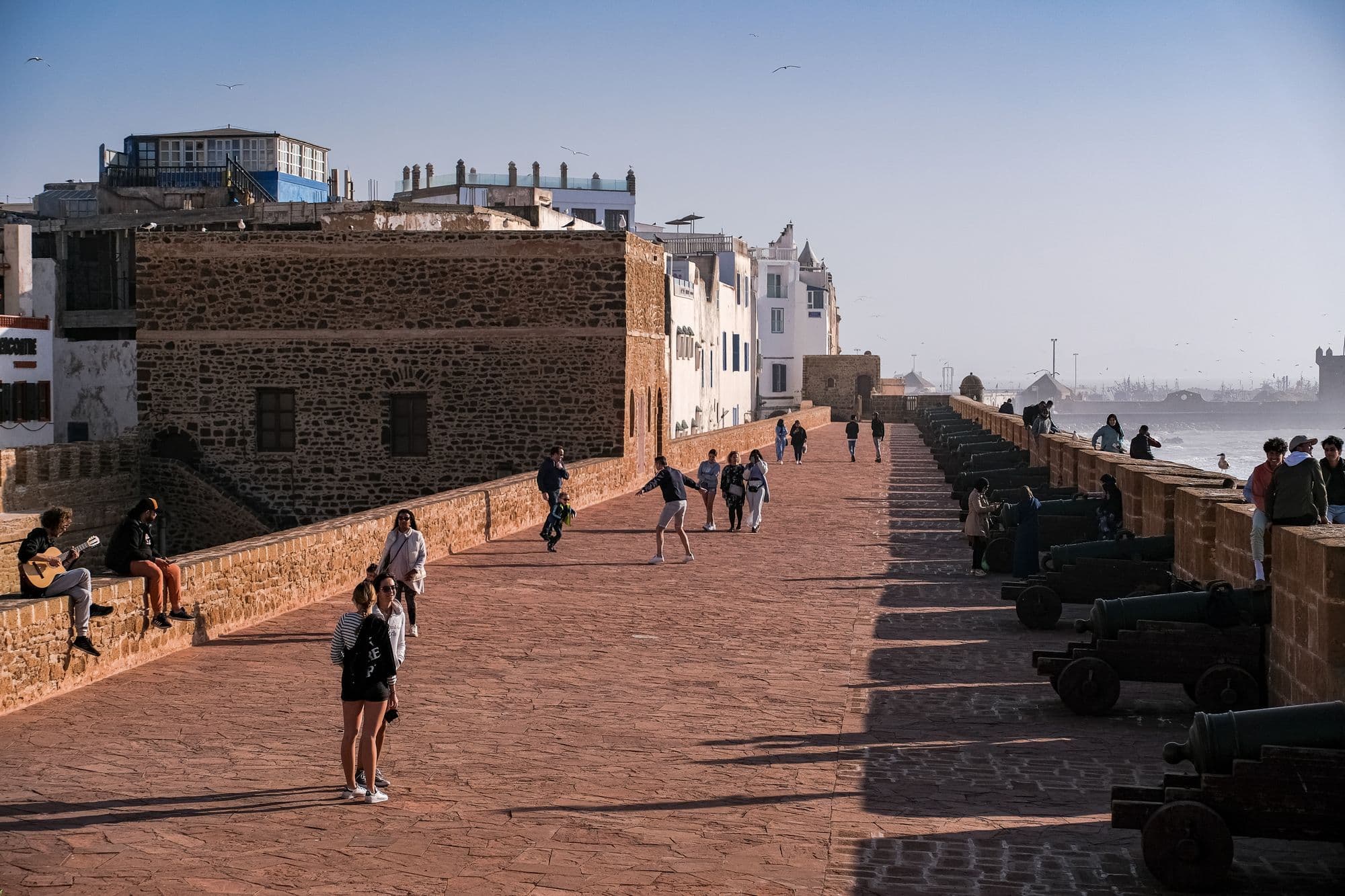 Visitors walk along the broad sea wall of Essaouira's Skala, lined with old cannons, beside a stone fort.