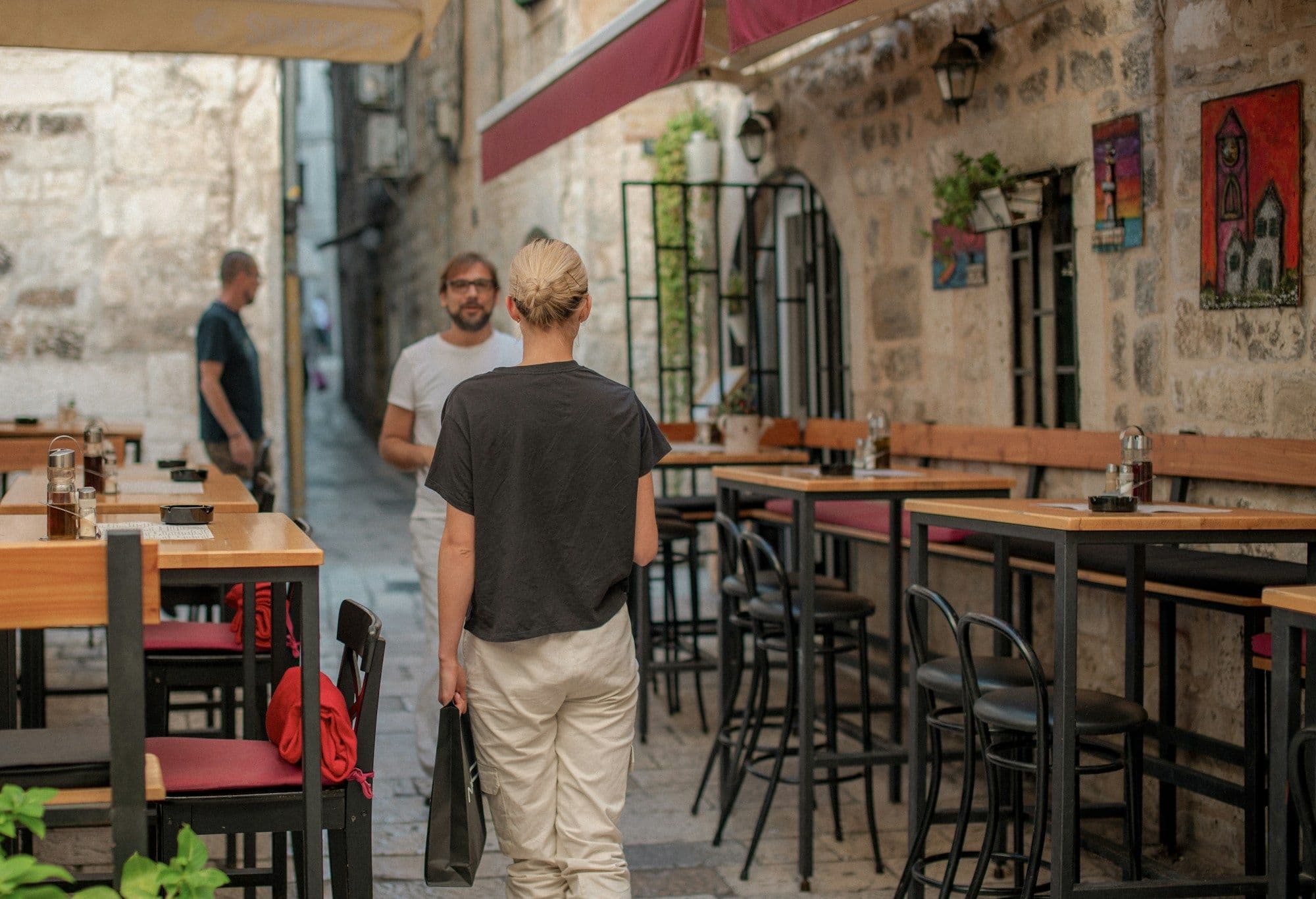 People walk past outdoor cafe tables on a narrow city street.