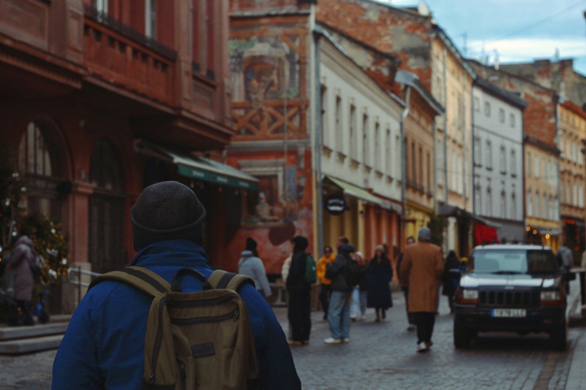 A traveler with a backpack seen from behind, walking down a cobbled old-town street lined with painted facades and cafés.