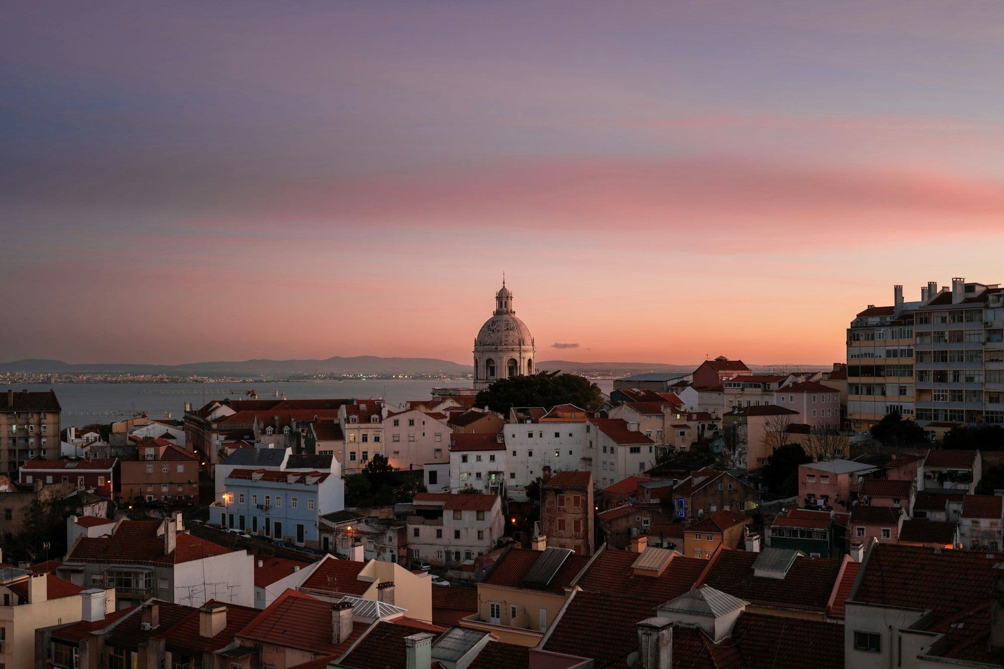 Terracotta rooftops at dusk with a white-domed church and the Tagus estuary under a pink and violet sky.