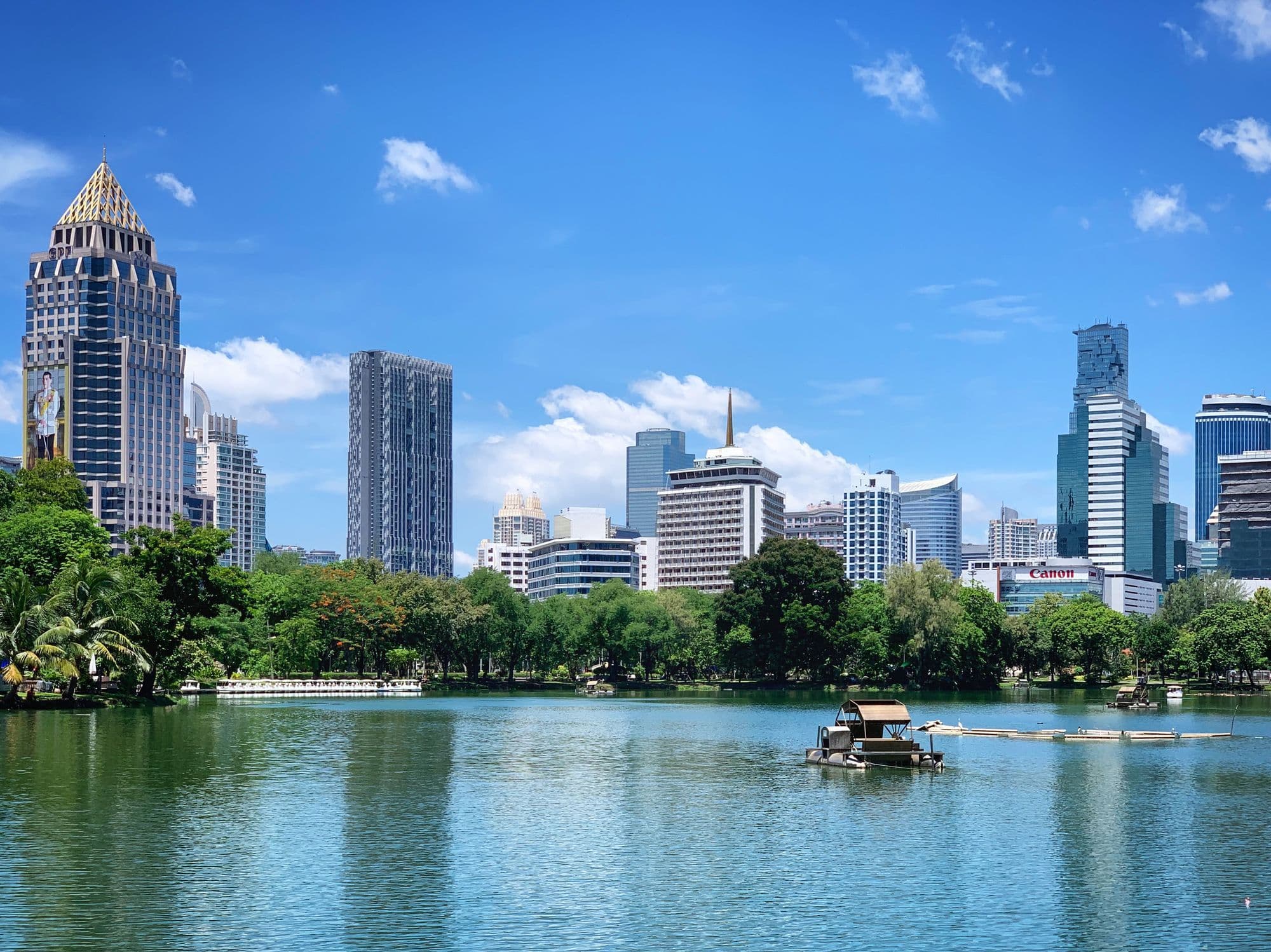 The Sathorn skyline reflected in the lake at Lumphini Park on a clear day, with a paddle boat in the foreground.