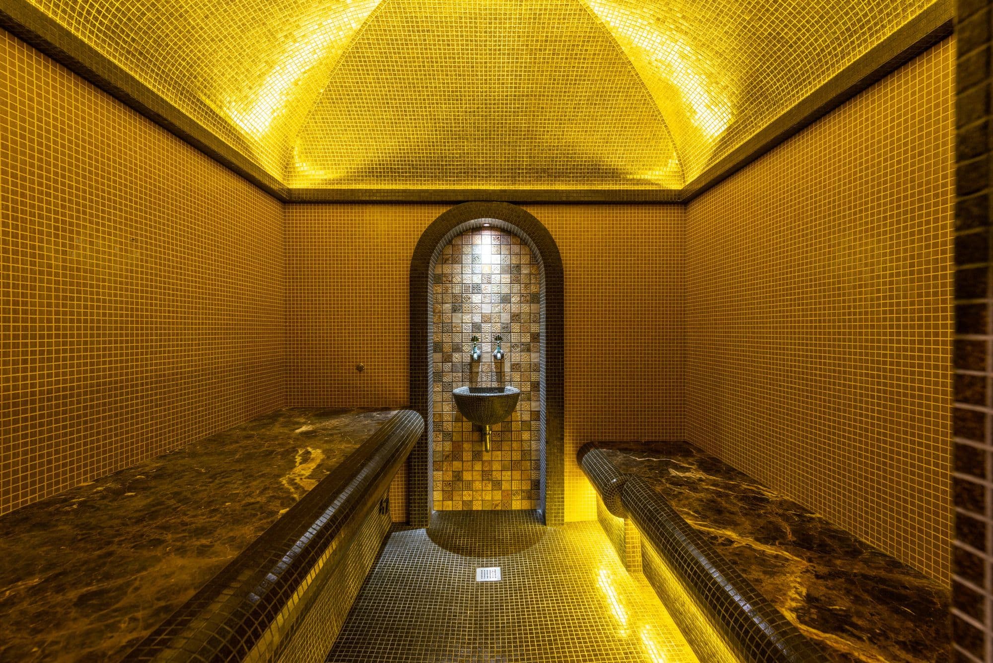 A domed steam room lined with gold mosaic tiles, with heated stone benches on both sides and a tiled niche at the end.