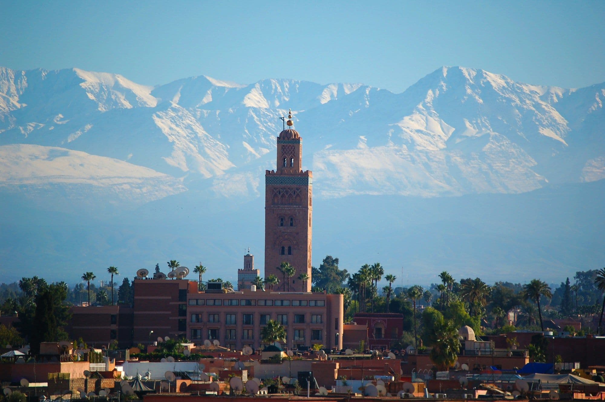 The Koutoubia Mosque minaret rising above Marrakech's rooftops, with the snow-capped Atlas Mountains behind.